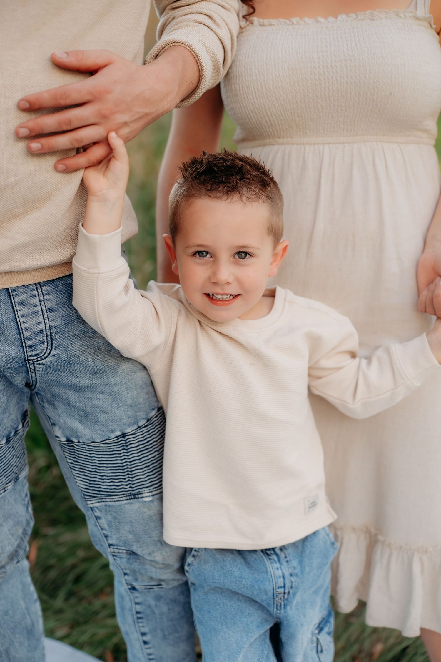 A smiling young boy in a cream sweater and jeans holds hands with his parents outdoors.