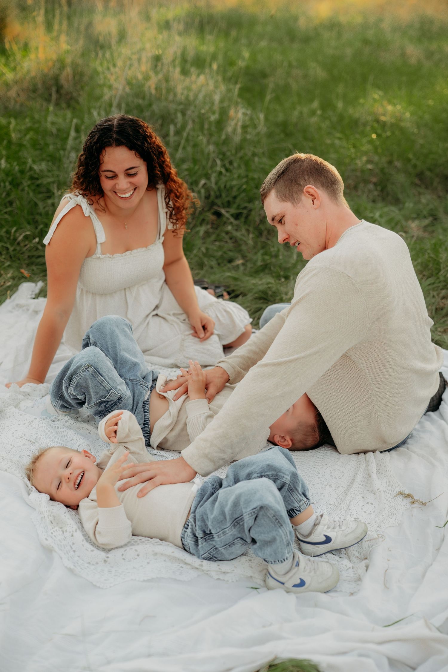 Happy family with a laughing baby on a white blanket in a green meadow during a sunny outdoor picnic.
