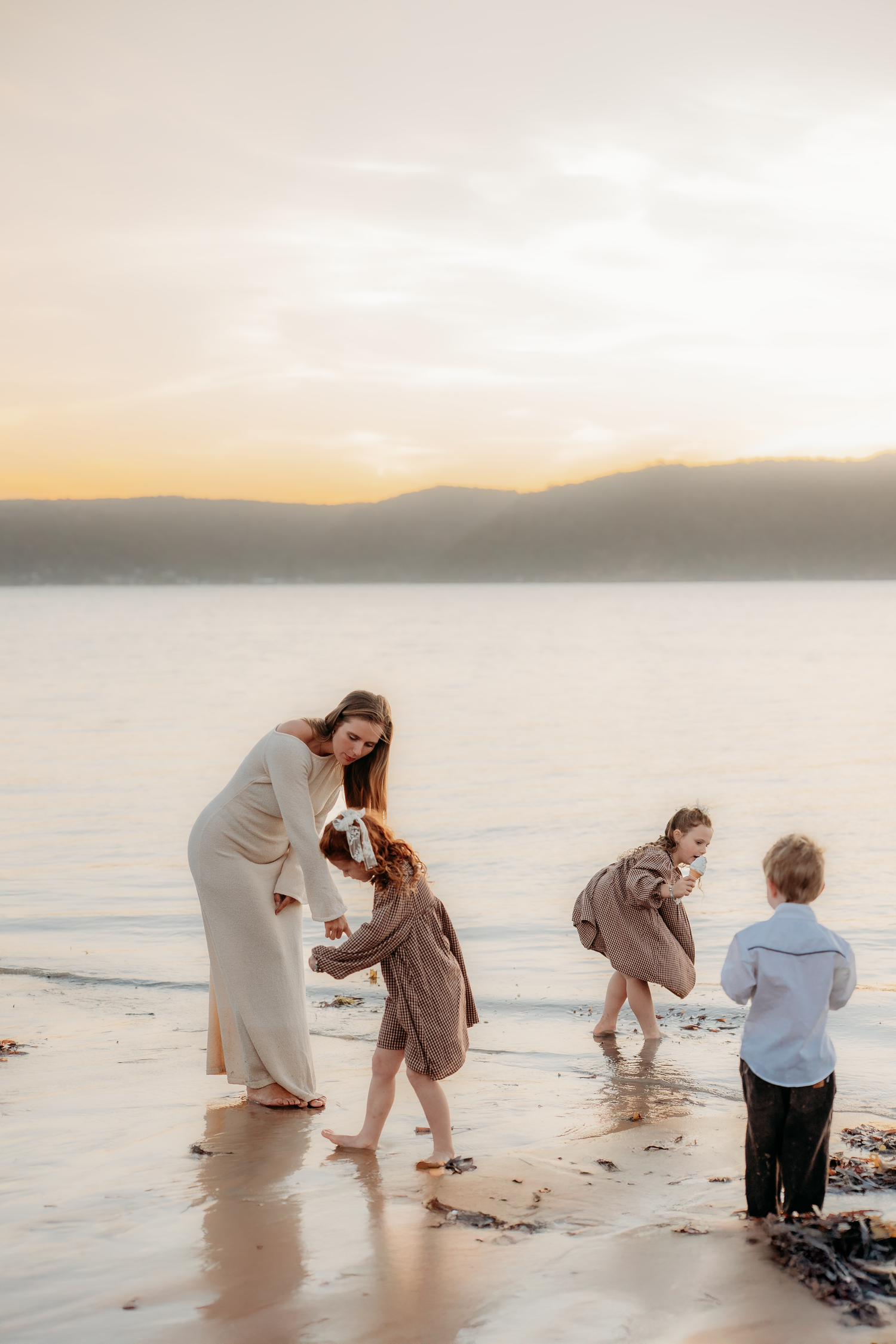 A mother and three children play on a sandy beach at sunset with mountains and calm water in the background.