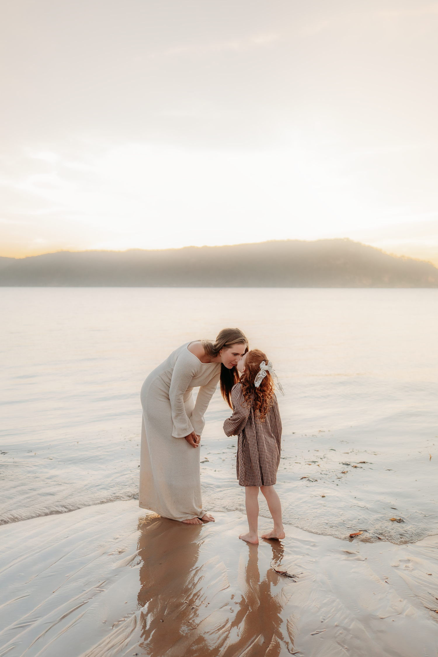 A mother tenderly bows to kiss her red-haired daughter while standing barefoot in shallow water at sunset.