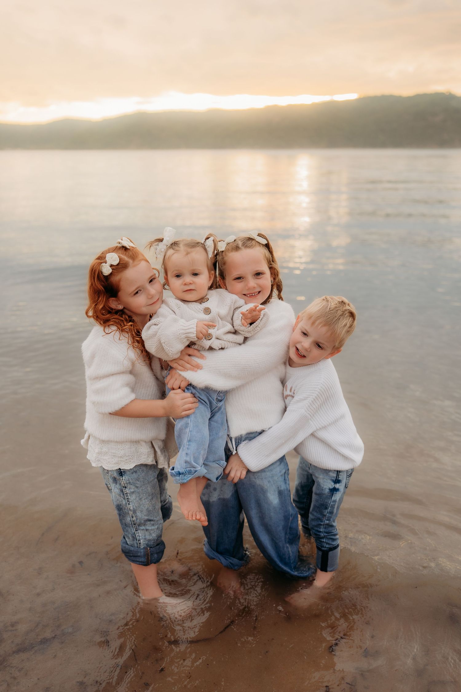 Four young children hugging together on a beach at sunset, wearing white sweaters and jeans.