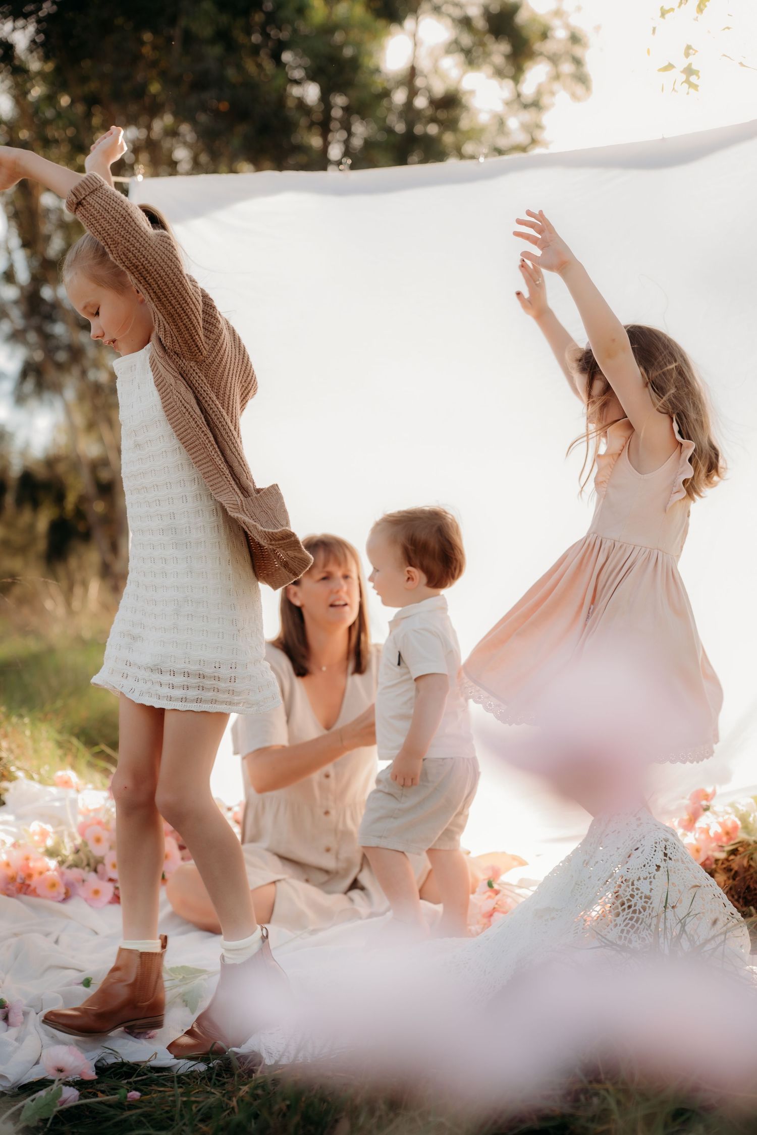 A mother sits on a white blanket outdoors while two young girls dance joyfully surrounded by pink flowers in soft sunlight.