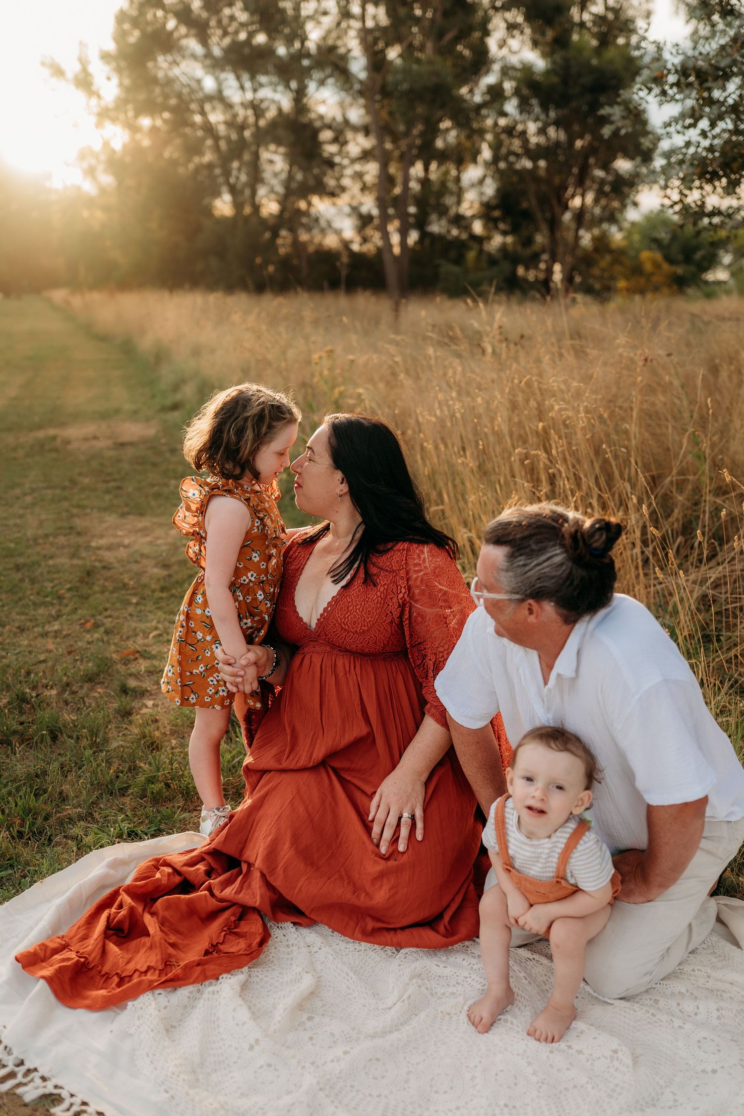 Family of four enjoying golden hour in a field, mother in red dress sharing a tender moment with toddler daughter.