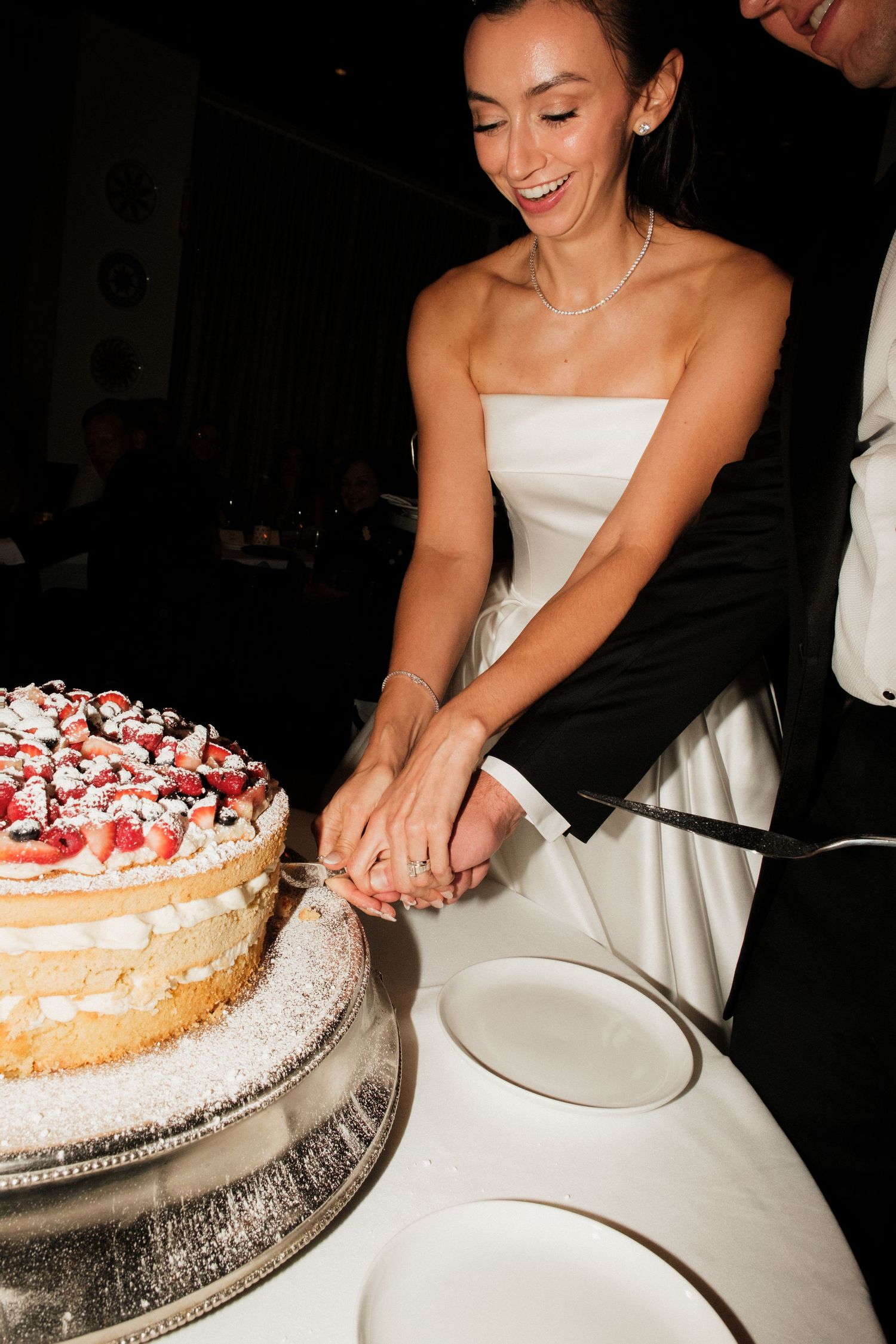 Smiling bride in white strapless gown cutting wedding cake topped with fresh berries with groom in black suit.