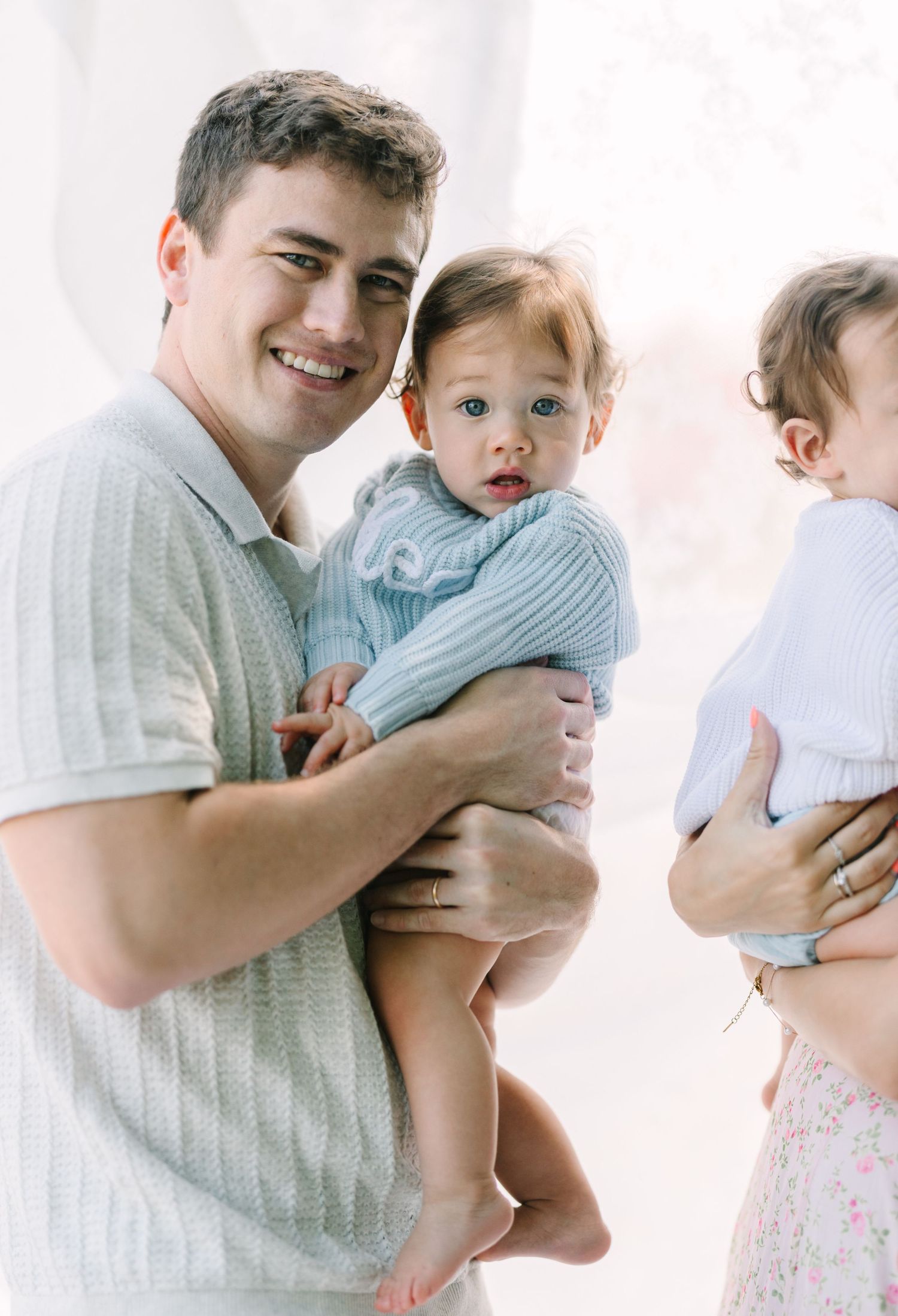 Smiling man holding a baby in a blue knit outfit, standing next to a woman holding another child indoors.