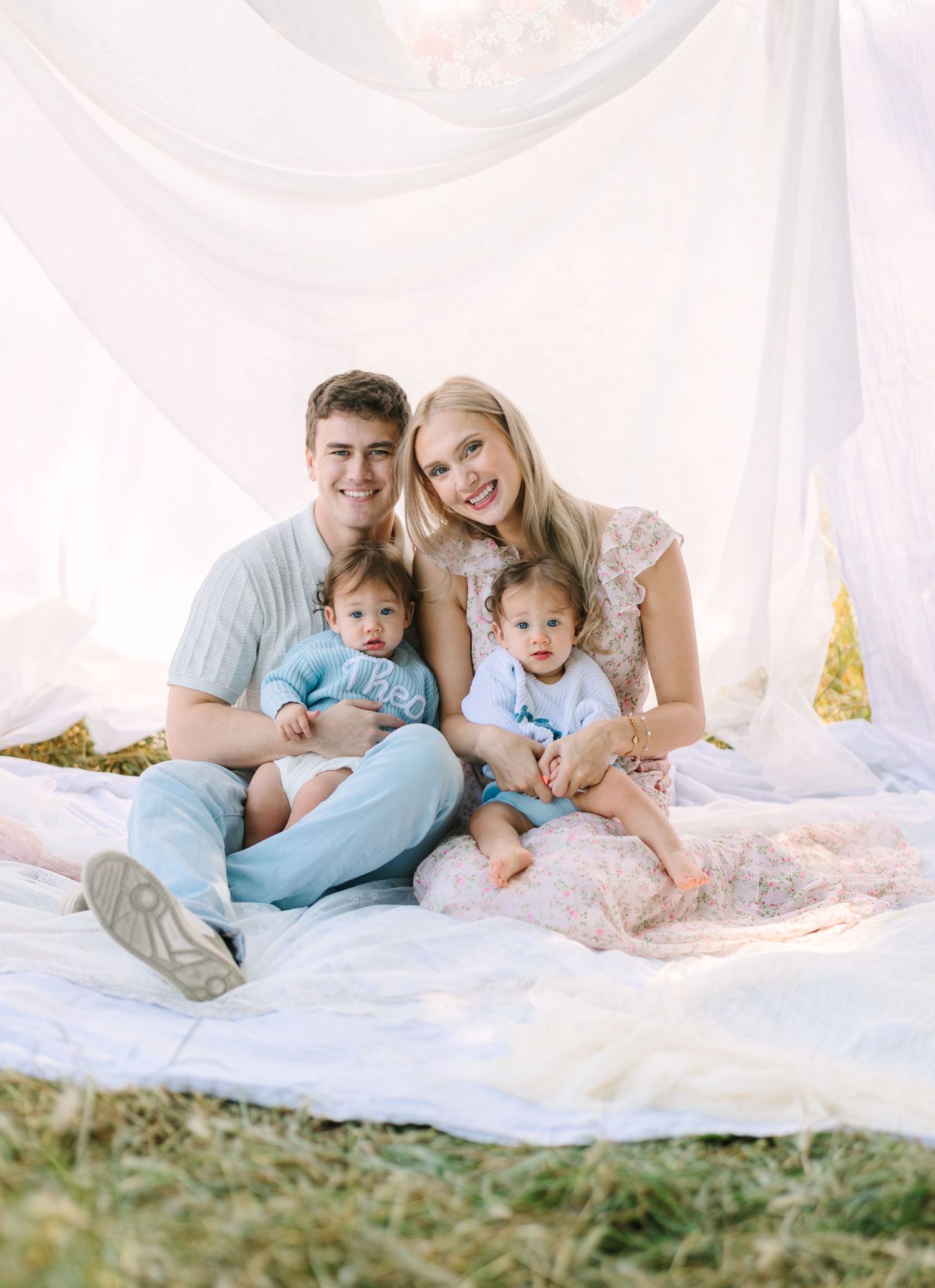 Young family with two babies sitting on a white blanket outdoors, smiling together in soft natural light.