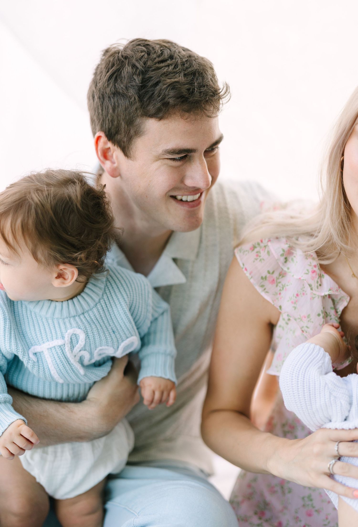 A smiling young man holds a baby in a blue knit outfit while a blonde woman in a floral dress cradles an infant.
