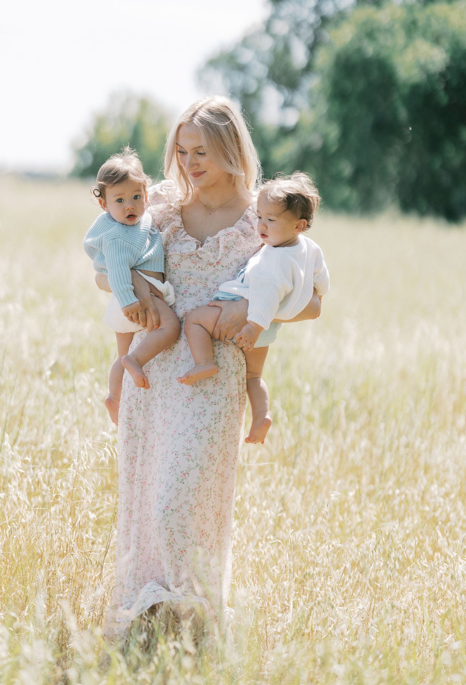 Blonde woman in floral maxi dress holding two toddlers in a sunny meadow field.