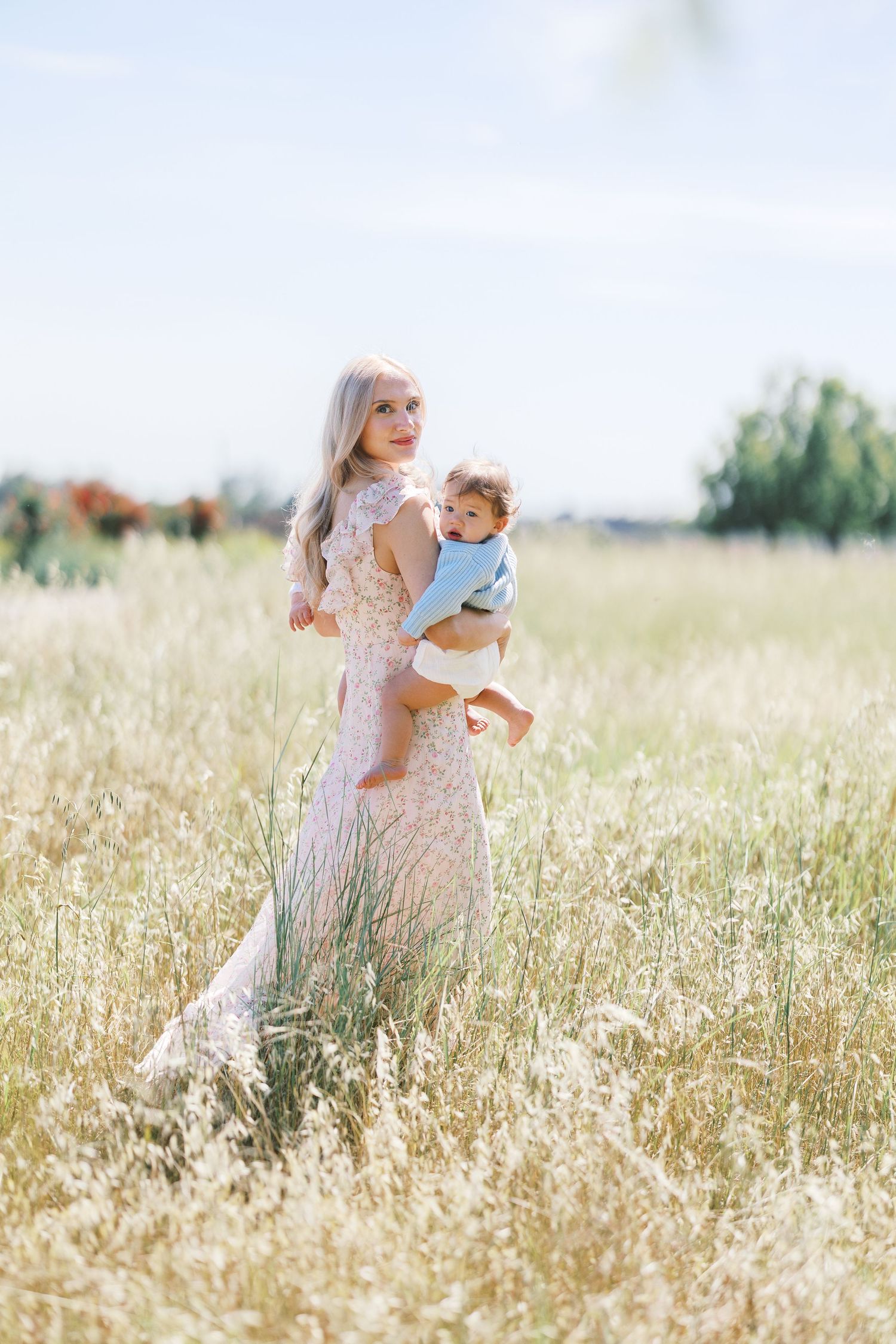 Blonde woman in lace dress carrying a baby through a sunlit grassy field.