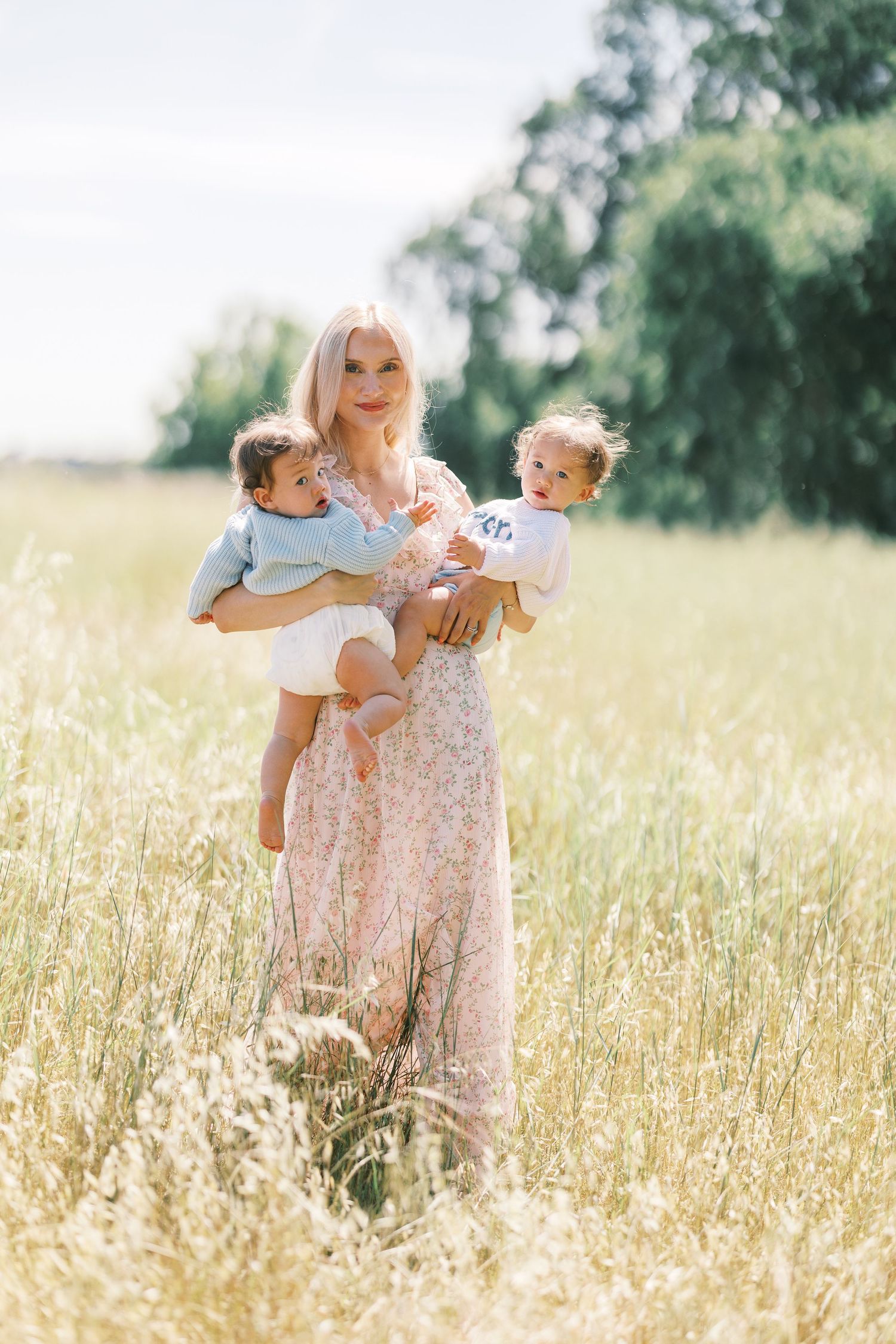 Blonde mother in floral dress holding two babies in a sunny golden field.