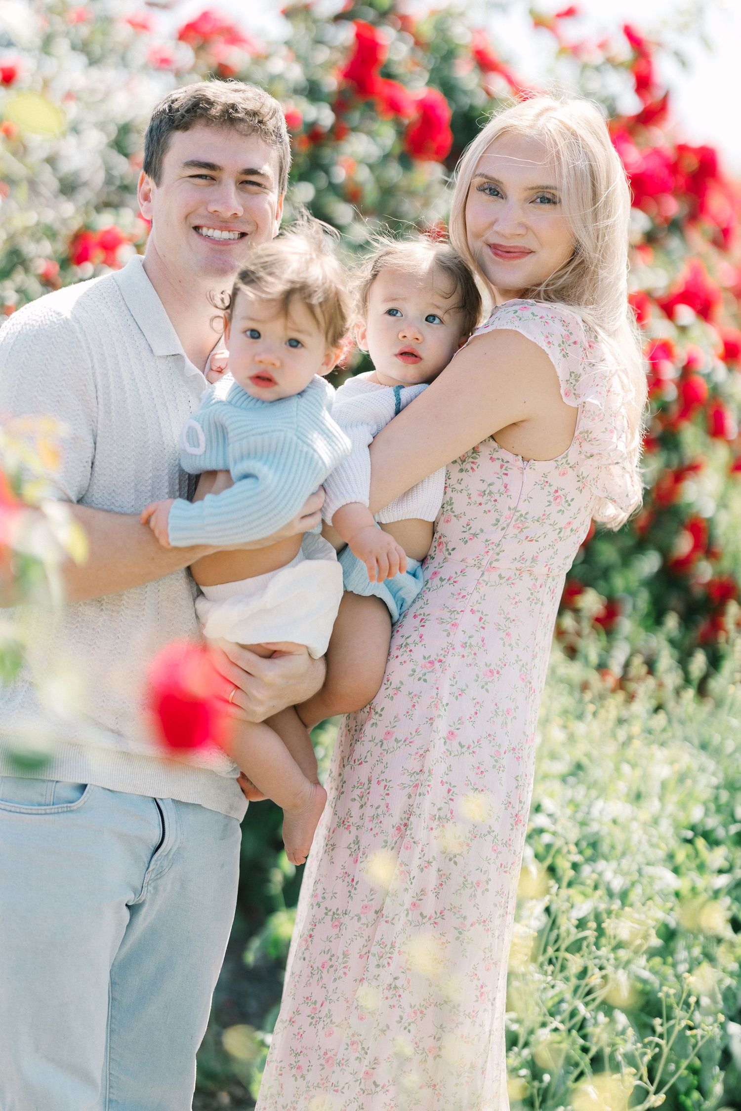 Happy family with blonde mother and father holding twin babies near red roses.