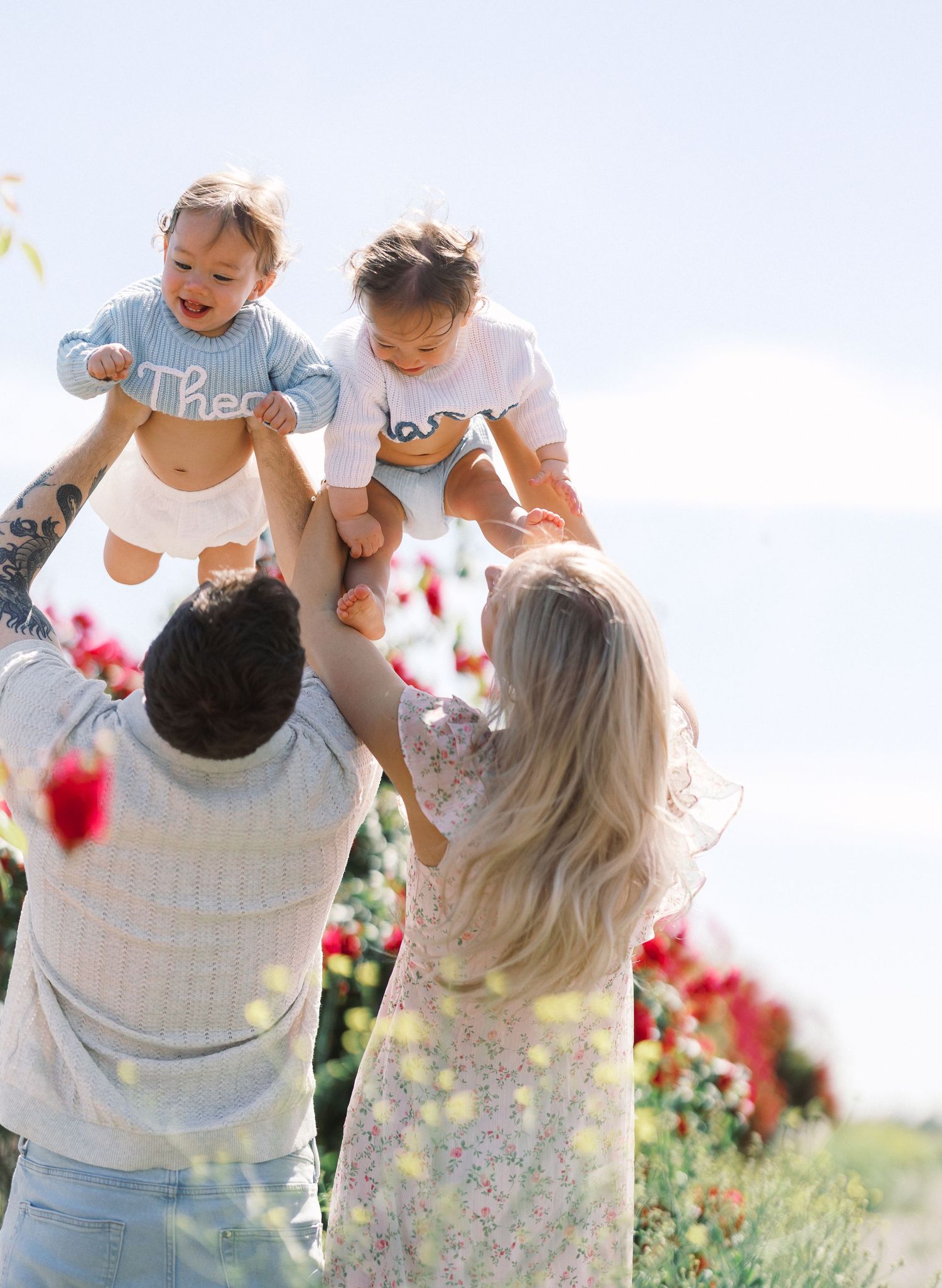 Parents lifting twin babies in the air in a colorful flower field on a sunny day.