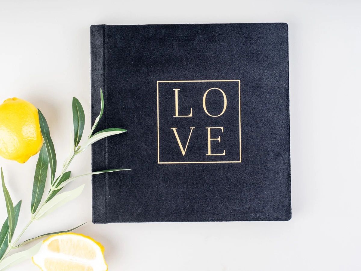 Black velvet photo album with gold LOVE text, displayed with fresh lemons and olive branches on white background.