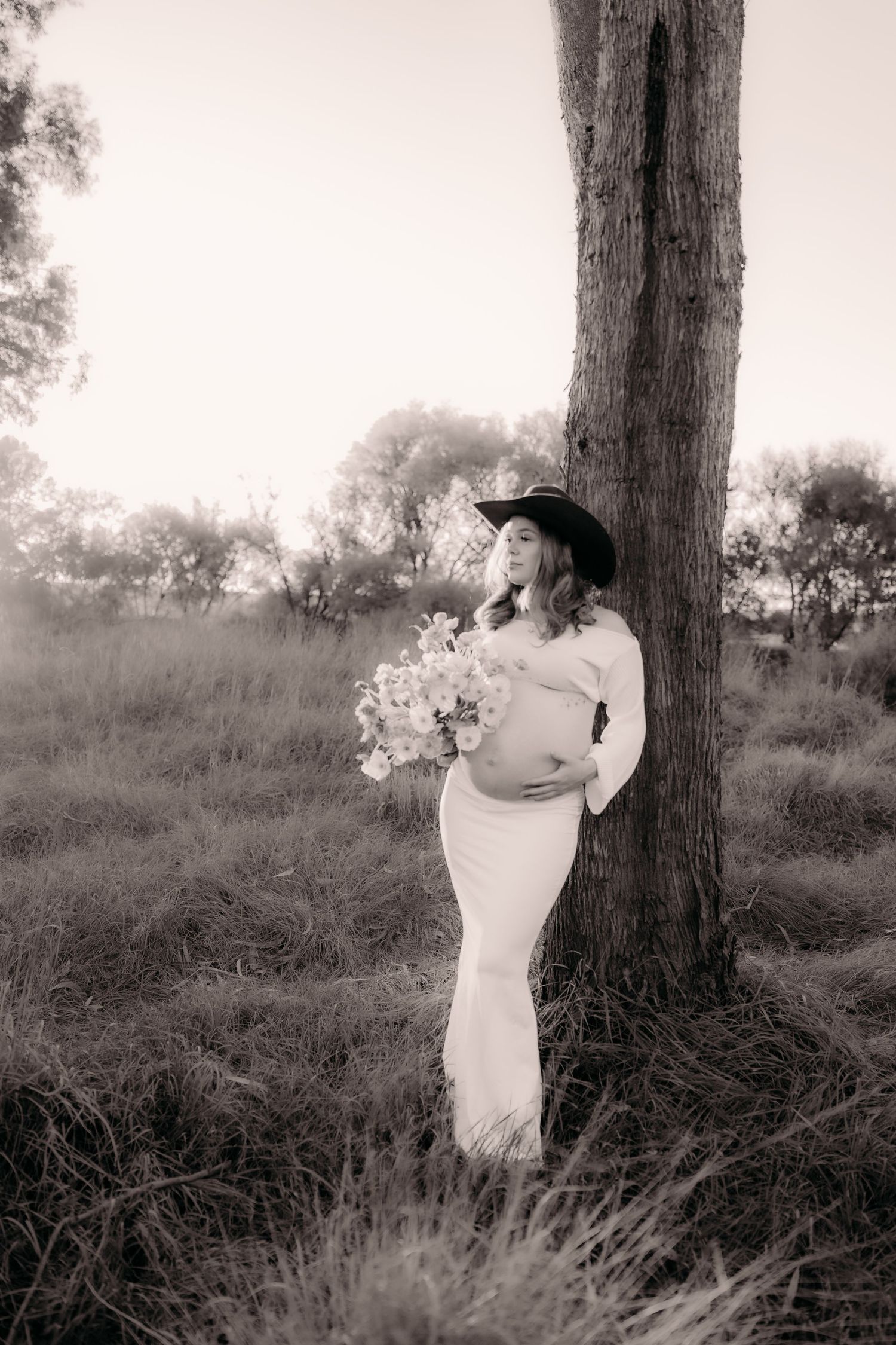 Pregnant woman in white dress and cowboy hat holds flowers beside a tree in a black and white outdoor maternity photo.
