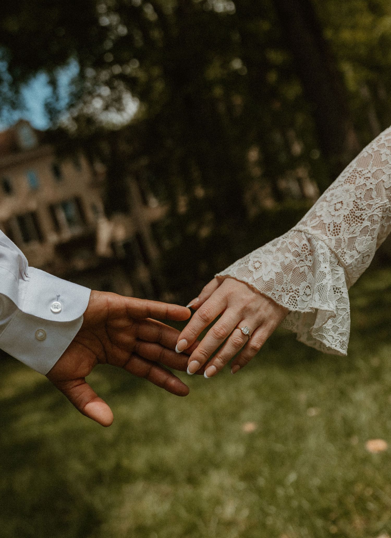 Couple holding hands outdoors, woman wearing lace wedding dress sleeve and engagement ring, man in white dress shirt.