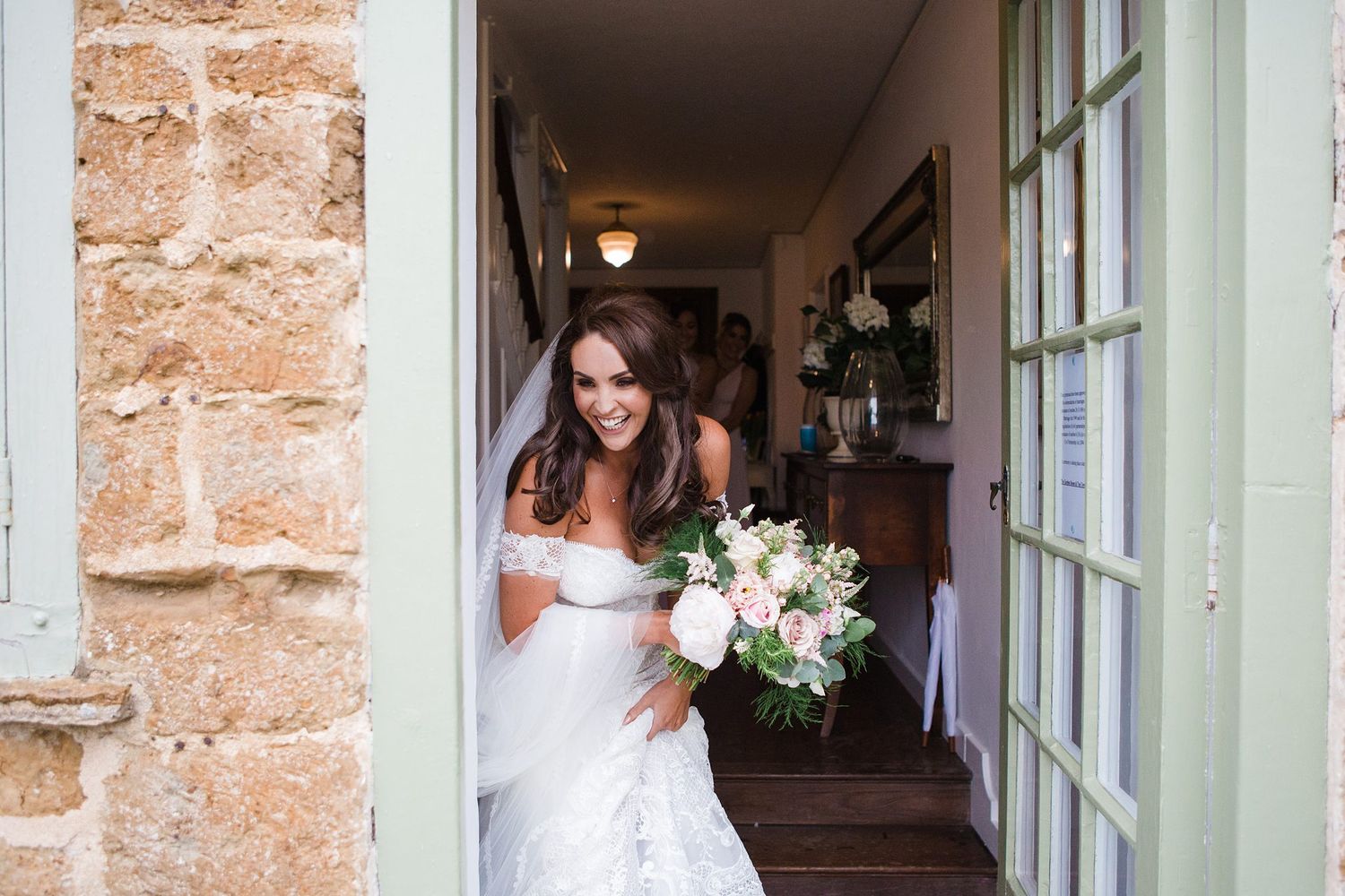 Smiling bride in white off-shoulder gown holding floral bouquet standing in doorway of rustic stone building.