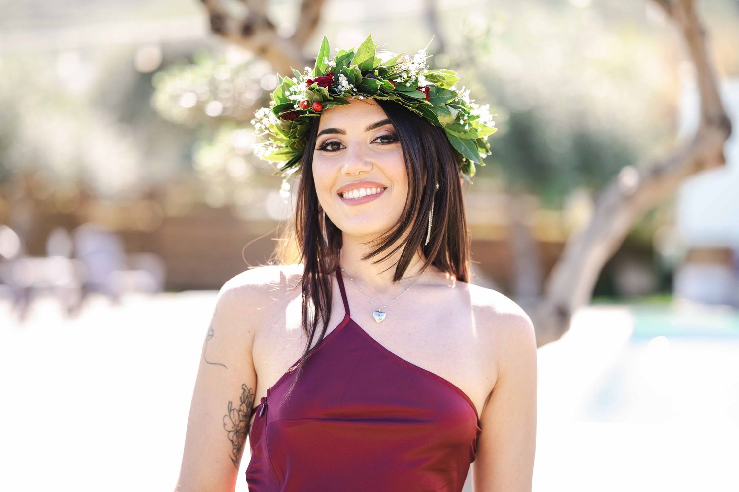 Donna sorridente con una corona di alloro e un tailleur bordeaux monospalla all'aperto durante una festa di laurea. Traguardo raggiunto.
