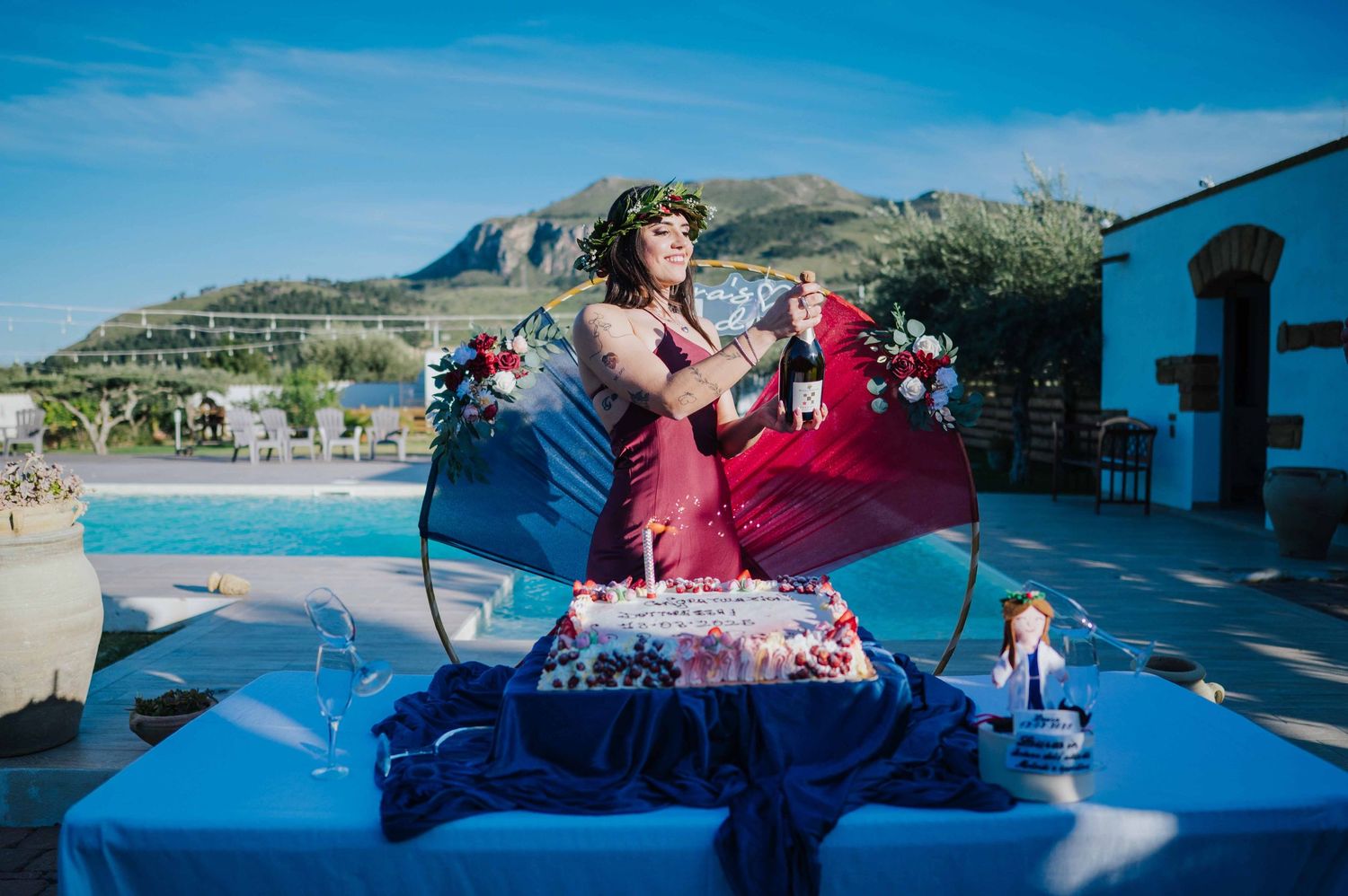 Dottoressa in abito rosso stappa una bottiglia di champagne accanto a una torta durante una festa a bordo piscina