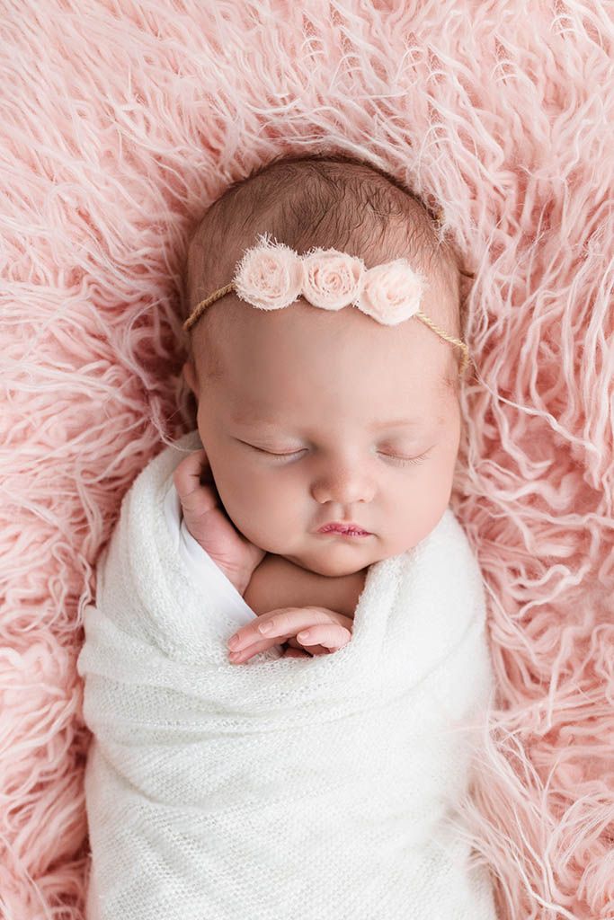 Sleeping newborn baby girl swaddled in white wrap with pink floral headband on pink fluffy blanket.