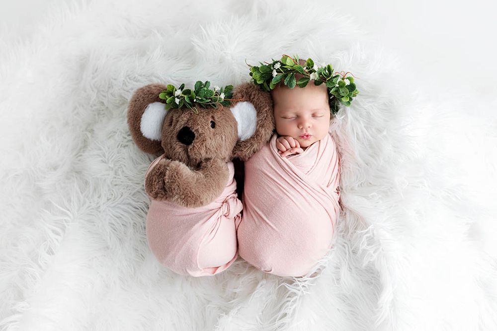 Newborn baby swaddled in pink wrap sleeping beside a koala stuffed animal, both wearing floral crowns on white fur.