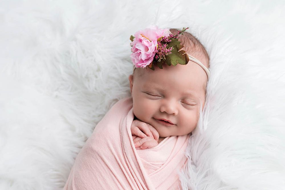 Smiling newborn baby girl swaddled in pink wrap with floral headband sleeping on white fluffy blanket.
