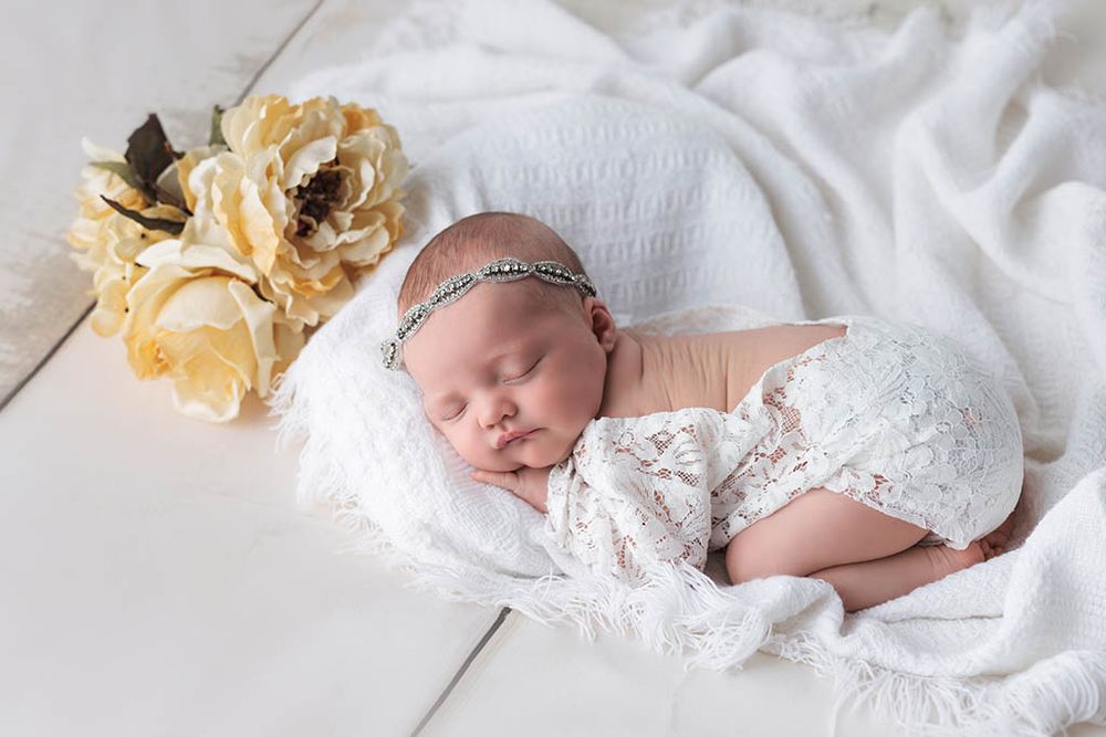 Sleeping newborn baby girl wrapped in white lace, wearing a delicate headband, posed with yellow flowers by Winchester CA baby photographer.