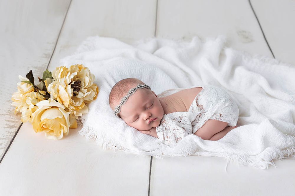 Peaceful newborn baby sleeping on white blanket beside yellow flowers in elegant lace wrap with headband by Winchester baby photographer.