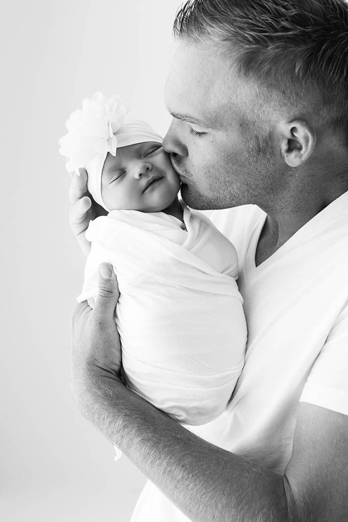 Dad kissing his newborn baby girl wearing white flower headband in black and white photo by affordable Winchester baby photographer.