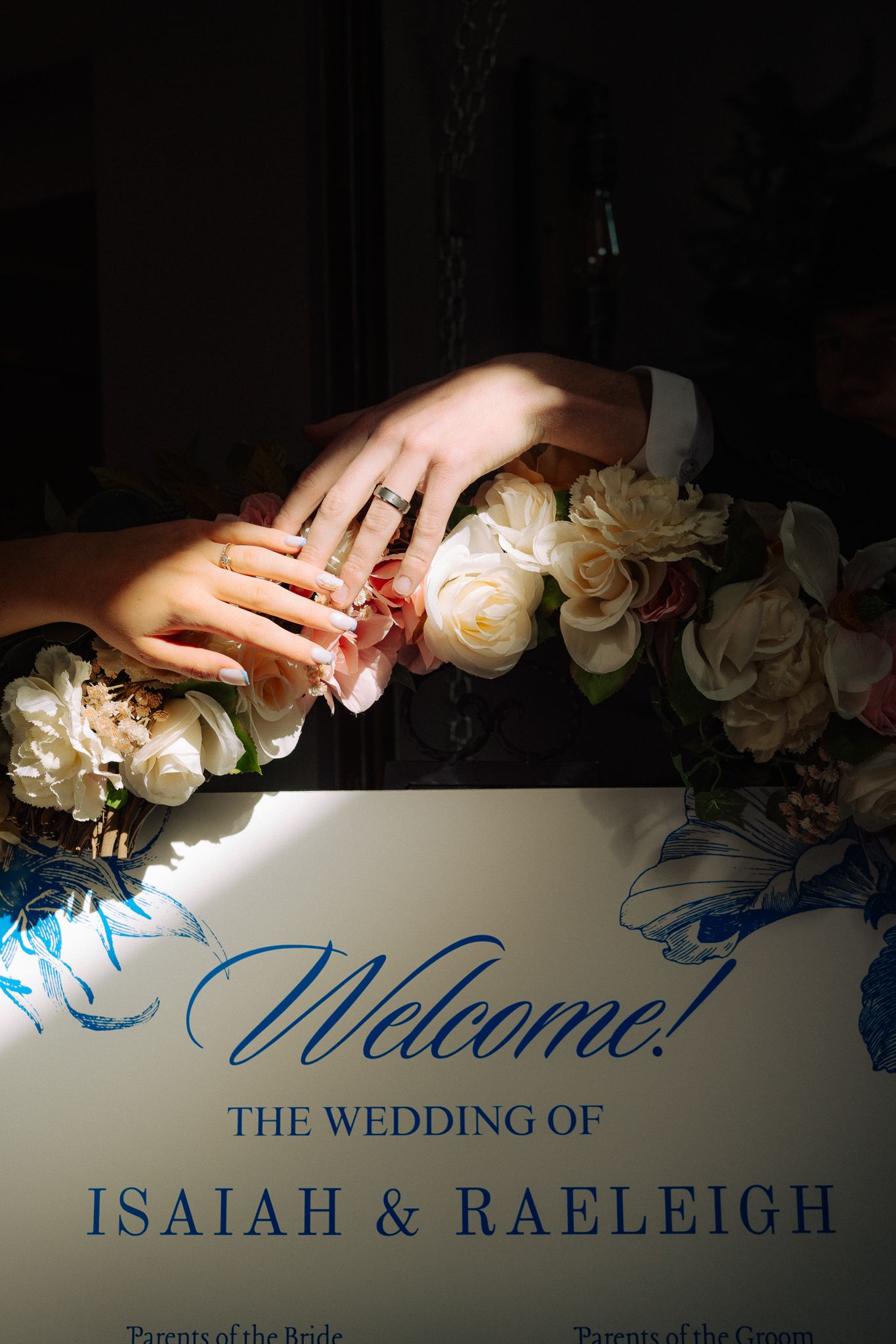 Newlywed couple's hands resting on a floral wedding welcome sign reading The Wedding of Isaiah and Raeleigh.