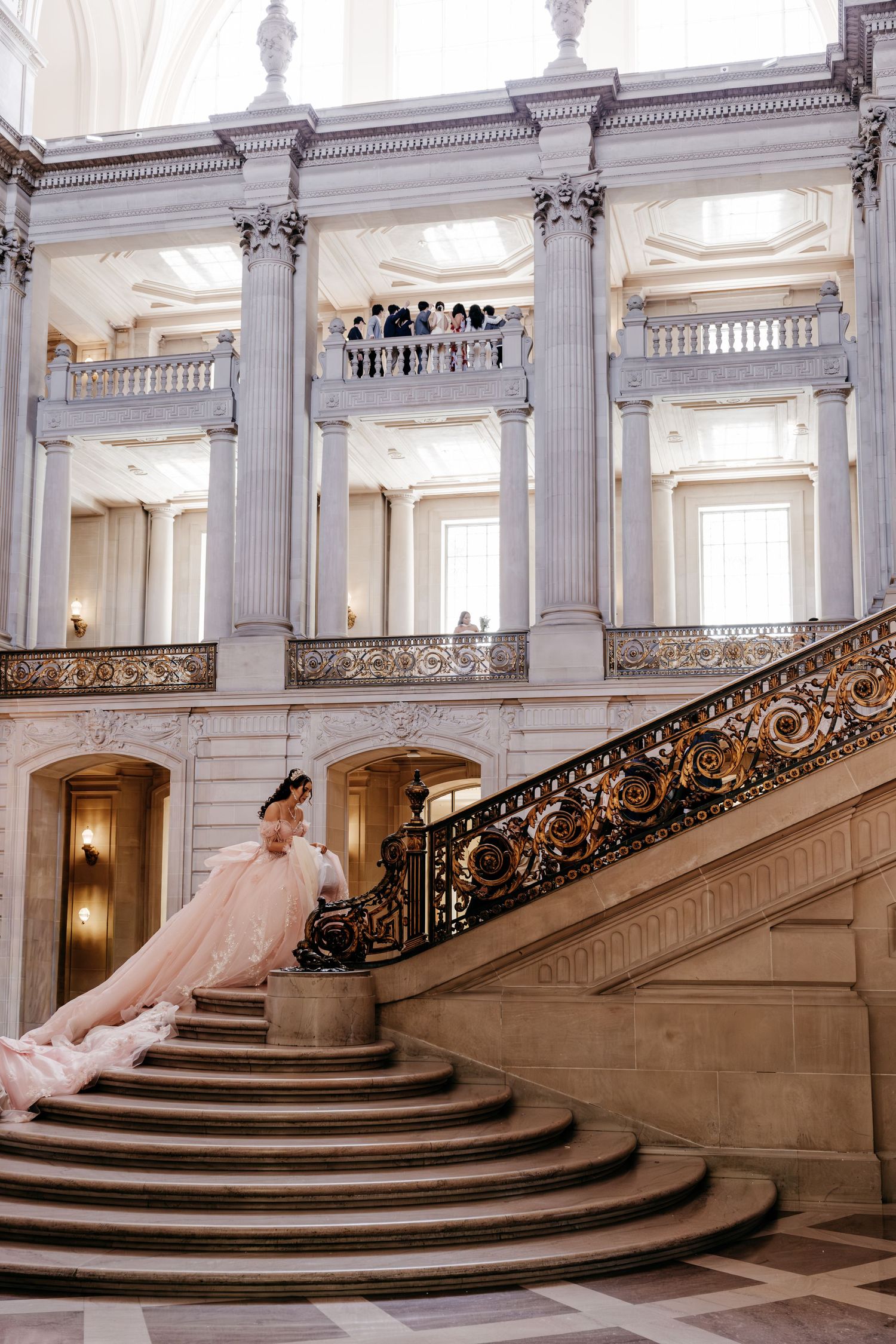 Bride in pink gown descends ornate staircase at San Francisco City Hall with grand columns and balcony.