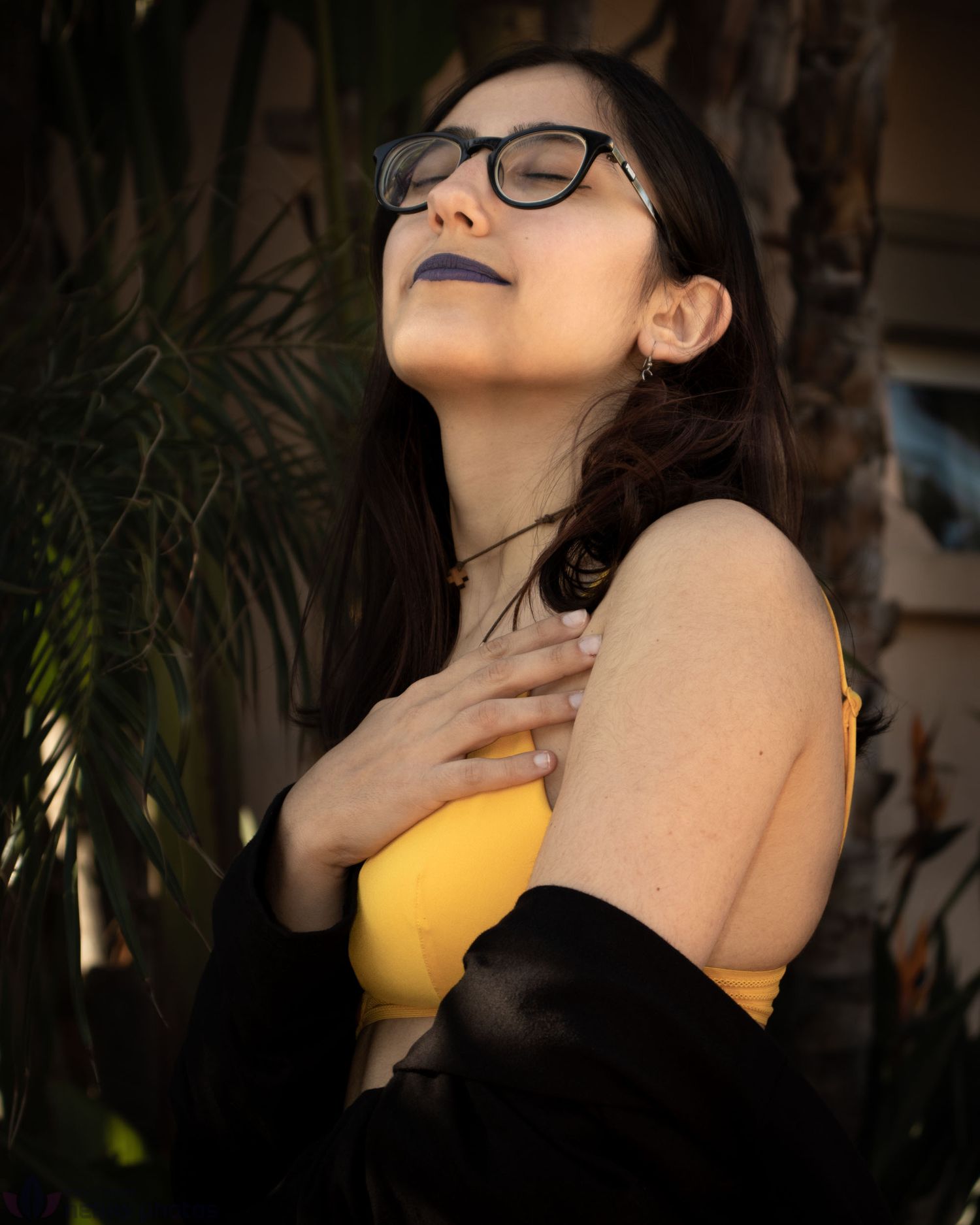 Young woman with glasses and dark lipstick, eyes closed, hand on chest, wearing yellow top outdoors.
