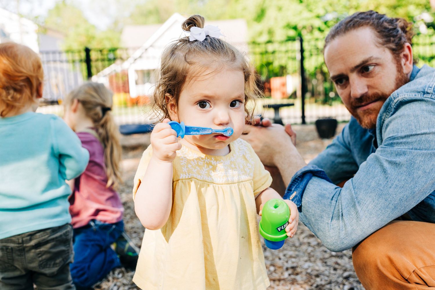 A toddler girl in a yellow dress blows bubbles outdoors while her father watches her at a playground.