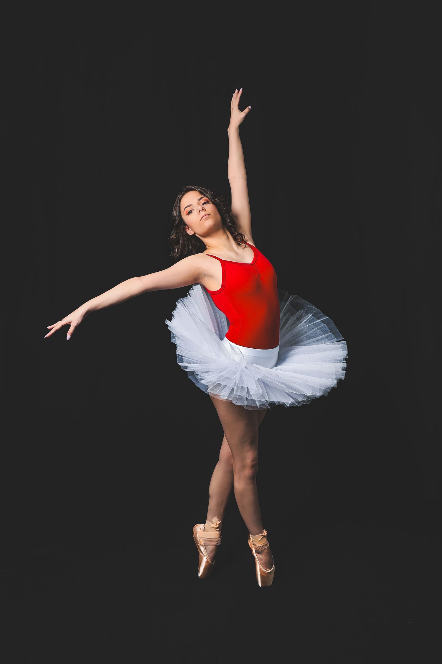 Ballerina in red leotard and white tutu performing graceful pose on pointe shoes against black background.