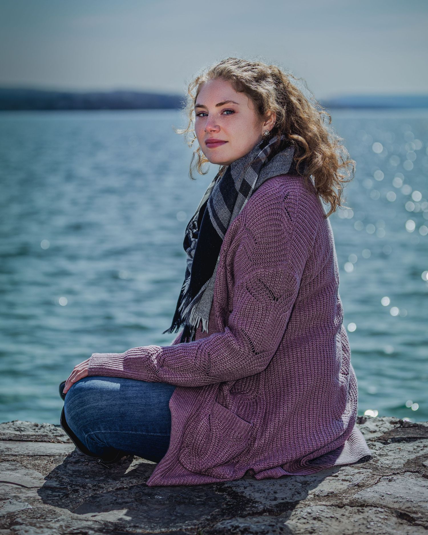 Young woman with curly hair sits on rocky shore wearing pink knit cardigan and scarf with blue water behind her.