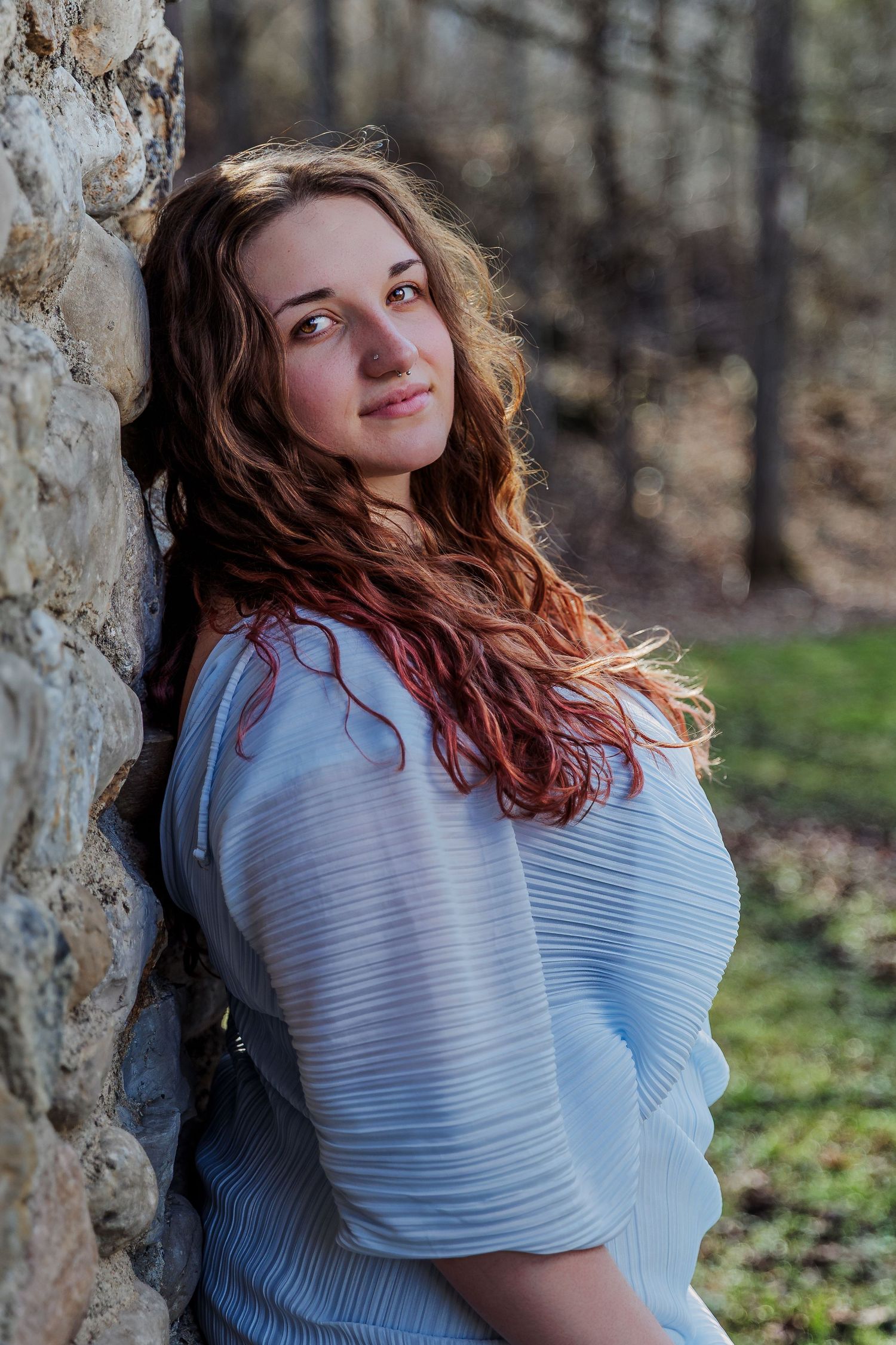 A young woman with long red wavy hair leans against a stone wall outdoors, wearing a light blue blouse.