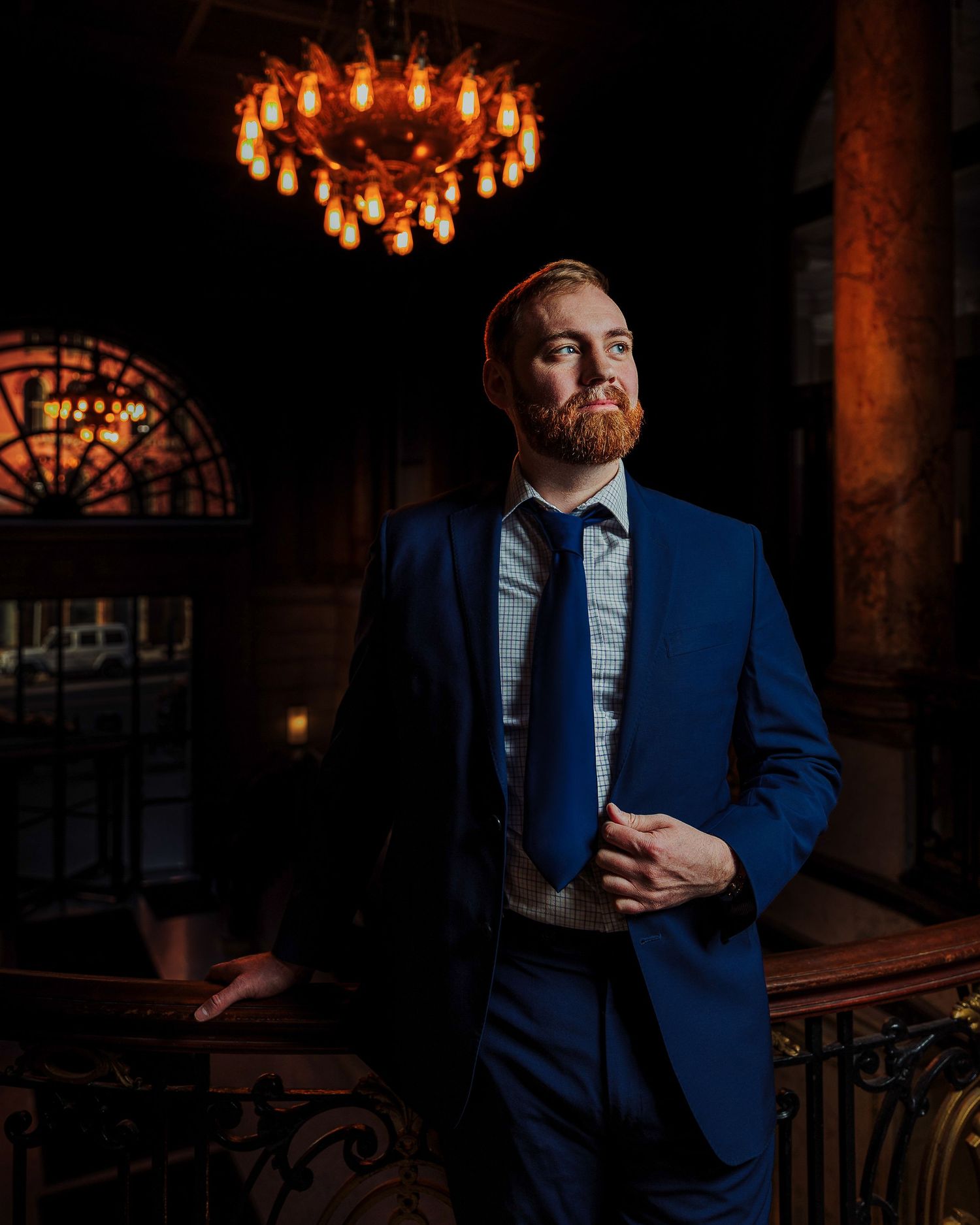 Man in blue suit with beard stands under chandelier in elegant venue.