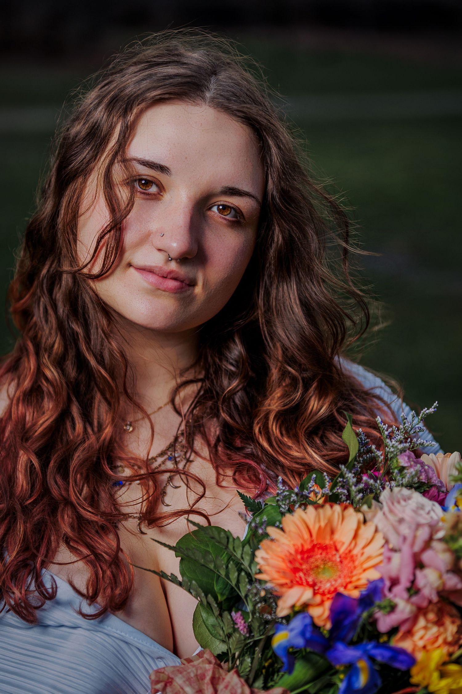 Young woman with curly auburn hair holding a colorful bouquet of flowers, posing outdoors in soft golden light.