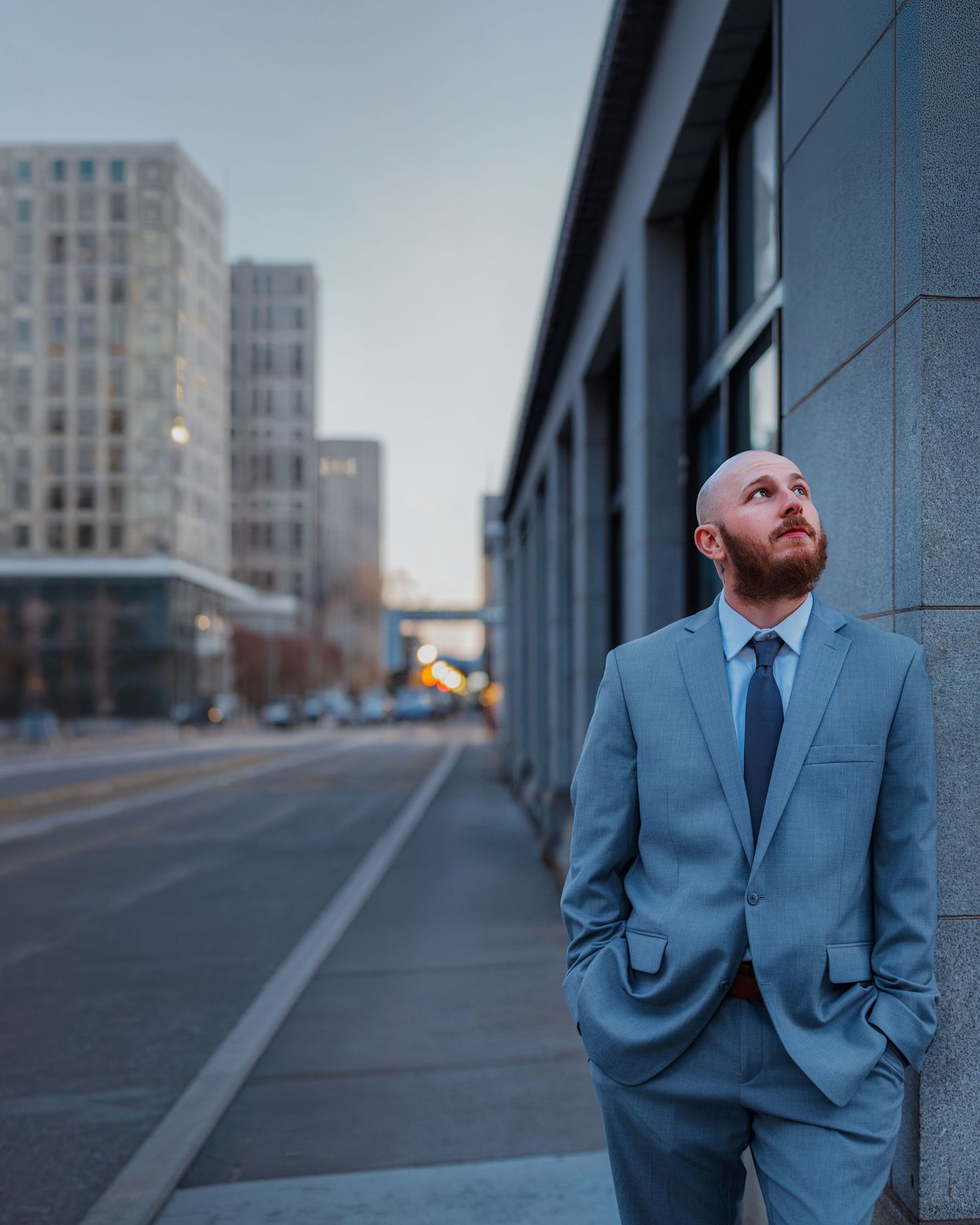 Bald bearded man in light blue suit leaning against urban building wall, looking up at city skyline at dusk.