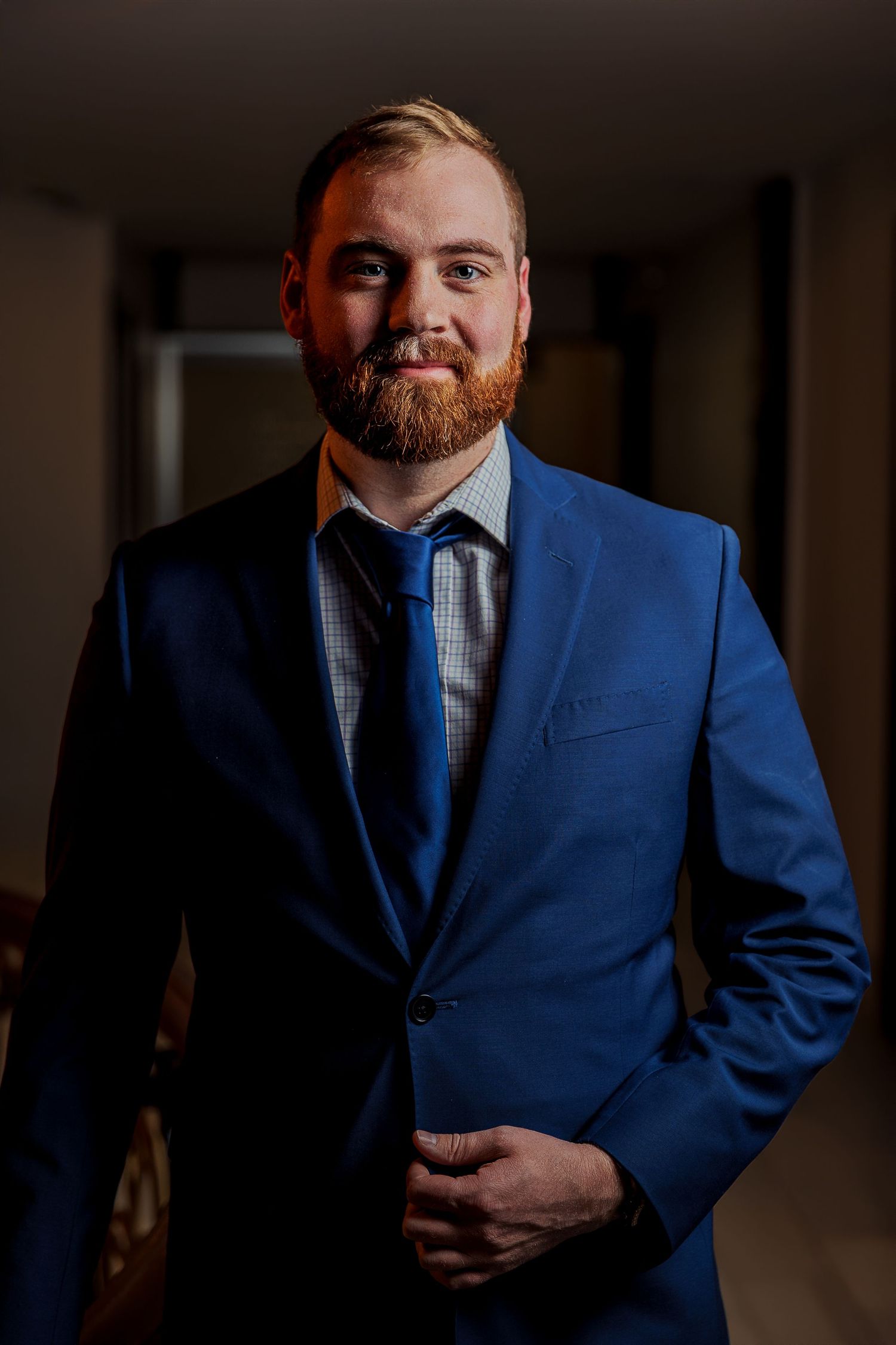 Man with red beard smiling in blue suit and tie, posing confidently indoors in formal attire.
