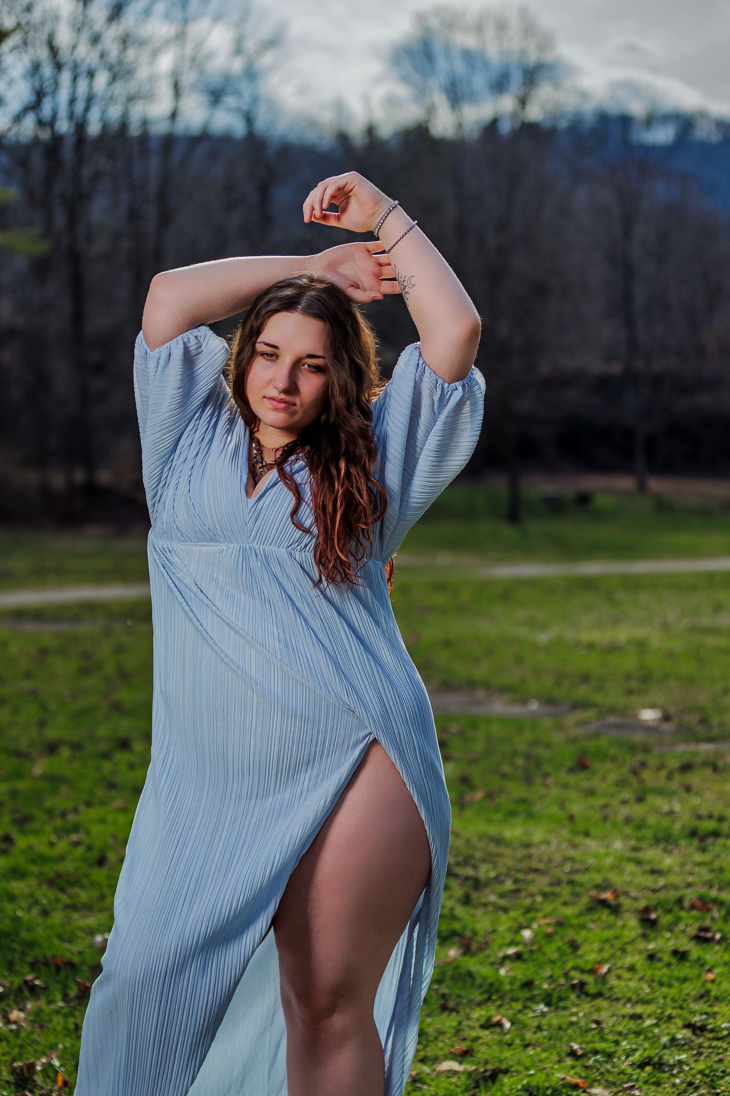 Young woman in light blue maxi dress with slit poses gracefully outdoors in a park with mountains in background.