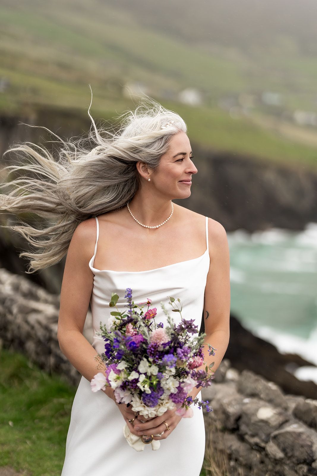 A bride in a white dress holds a vibrant purple and white bouquet on a windy cliffside overlooking the ocean.