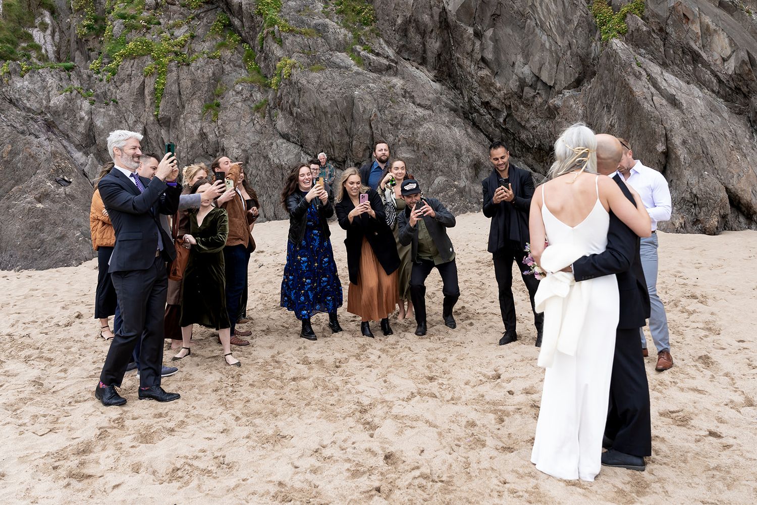 A group of people gather near rocky cliffs on a sandy beach during what appears to be a special occasion.