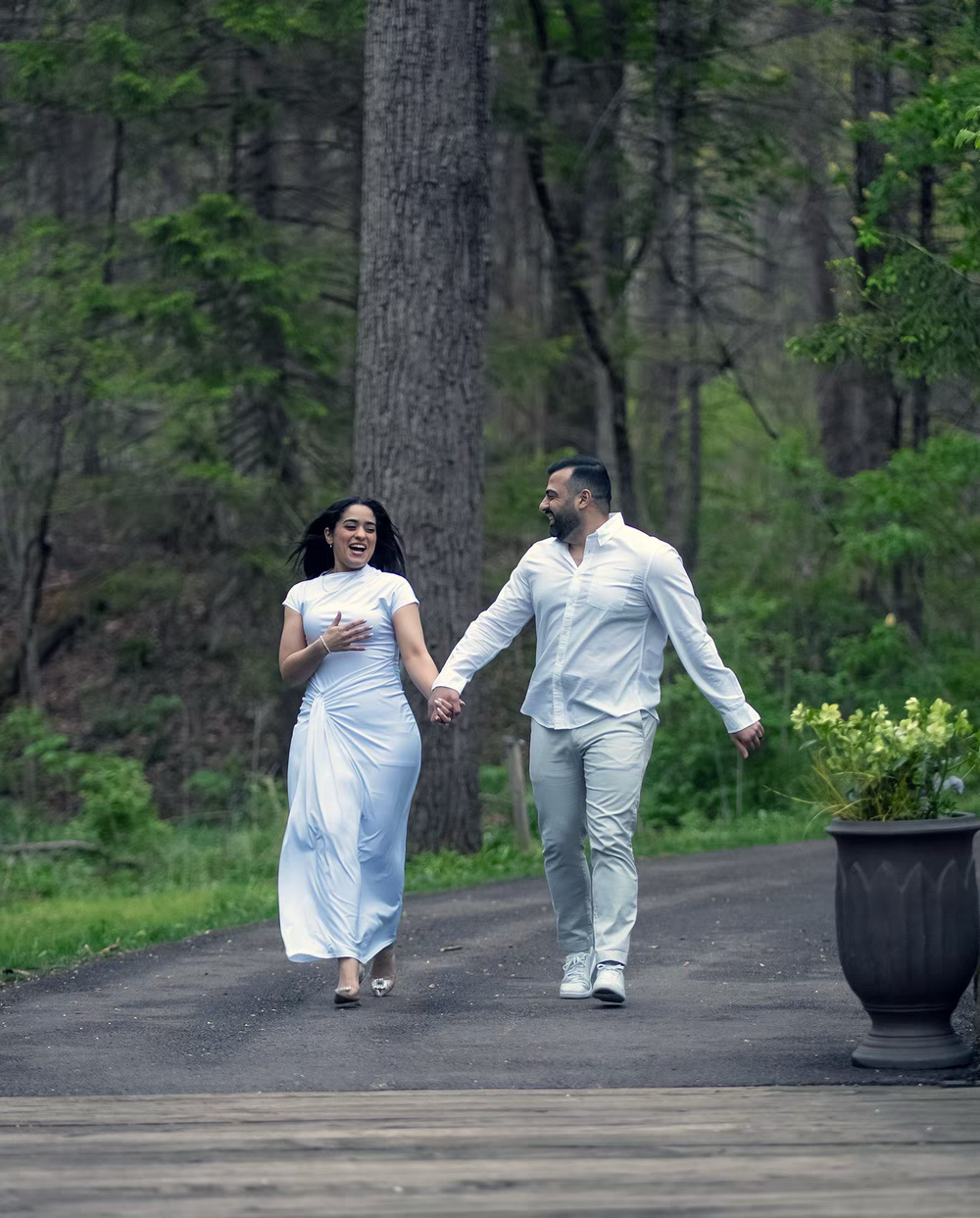 Couple in white outfits holding hands and walking on a path surrounded by lush green trees in a forest setting.