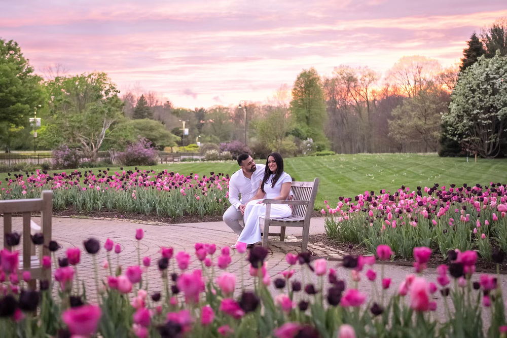 Couple in white sitting on bench surrounded by pink tulips at sunset in a lush garden.
