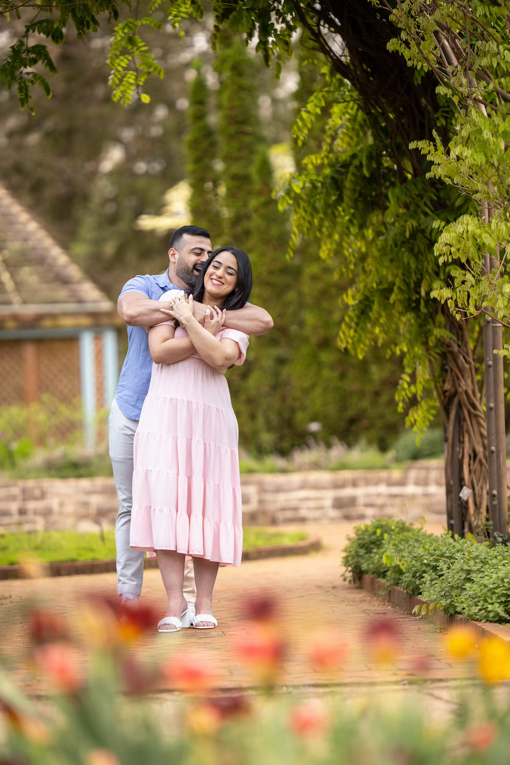 A couple embraces tenderly in a lush garden, the woman in a pink dress, surrounded by colorful tulips.