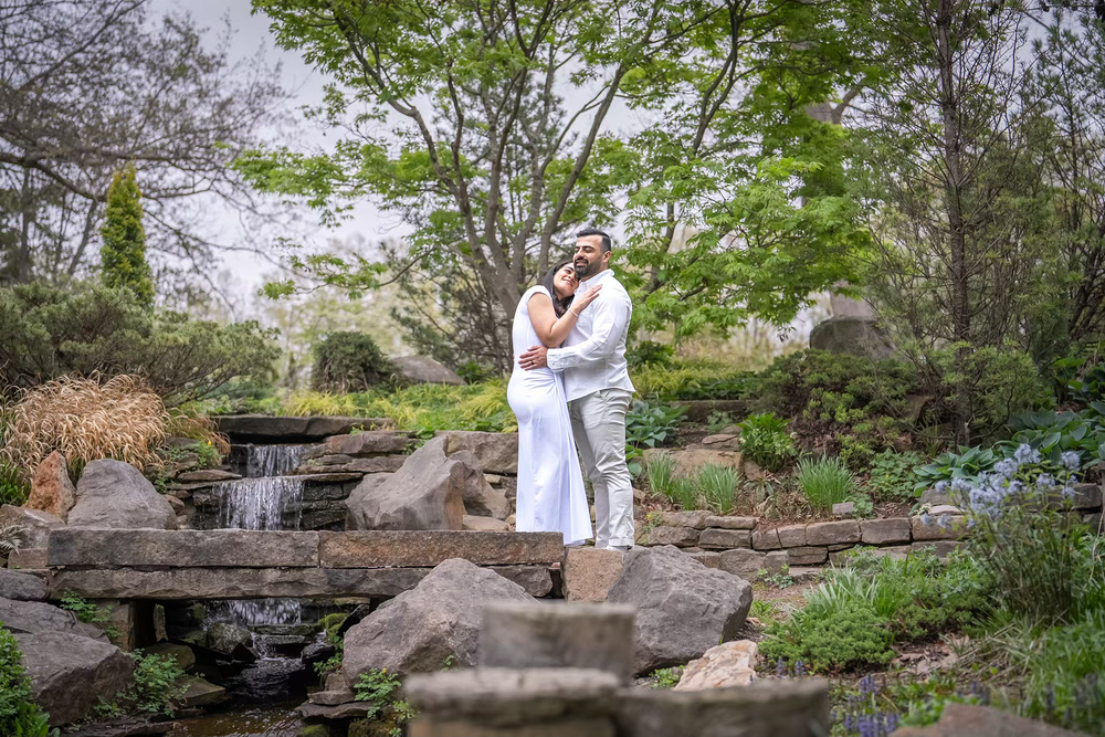 Couple embracing in white outfits on stone bridge surrounded by lush garden greenery and small waterfall.
