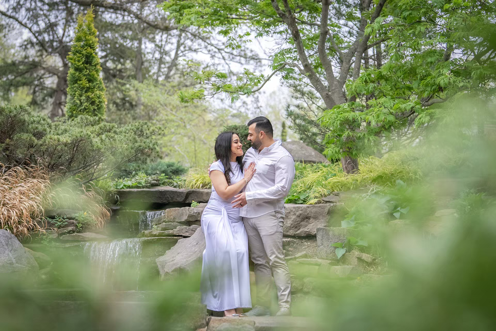 Couple in white embracing in a lush green garden with stone waterfall backdrop for engagement photos.