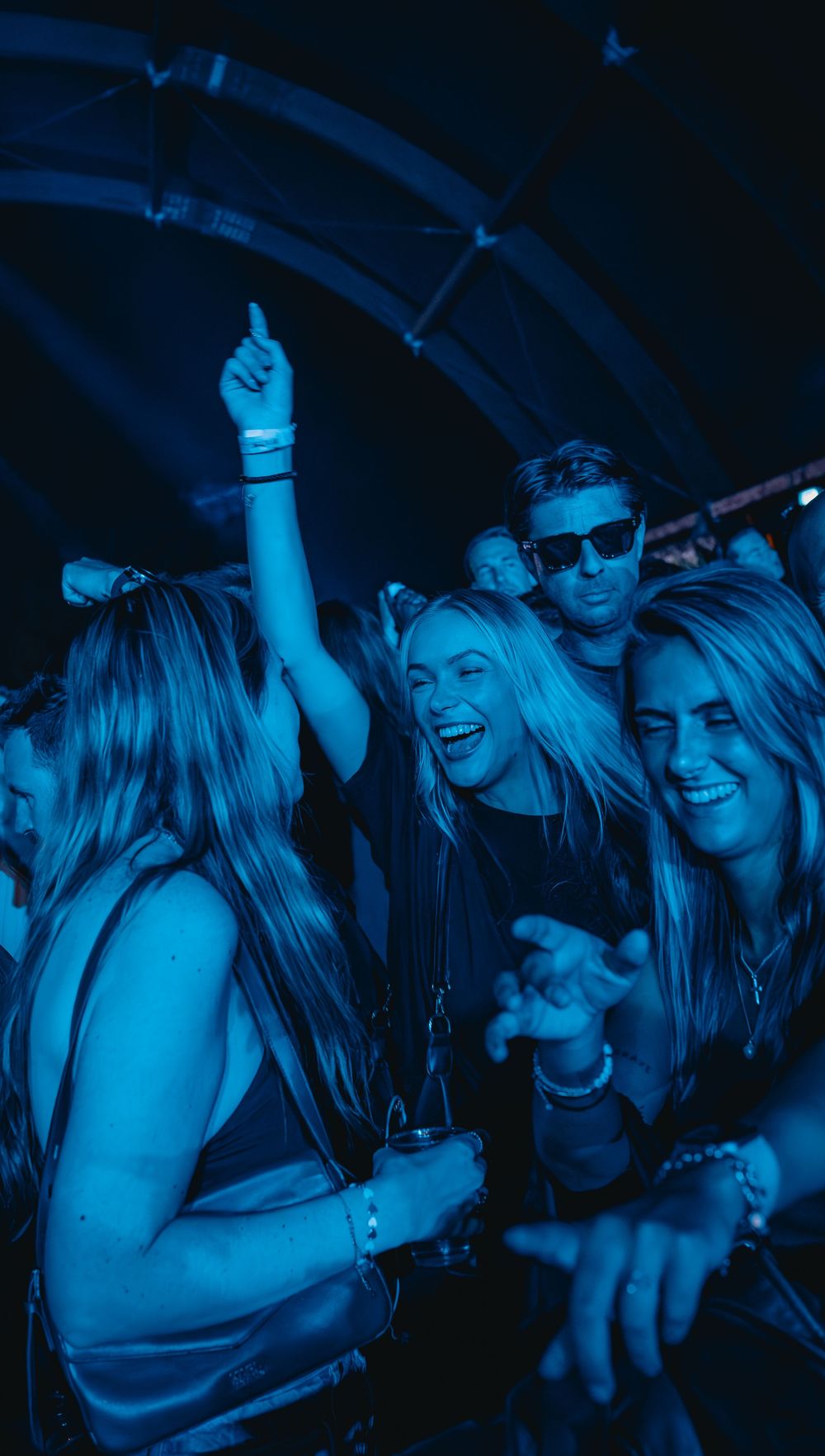 Excited crowd of young women dancing under blue lights at an outdoor music festival at night.