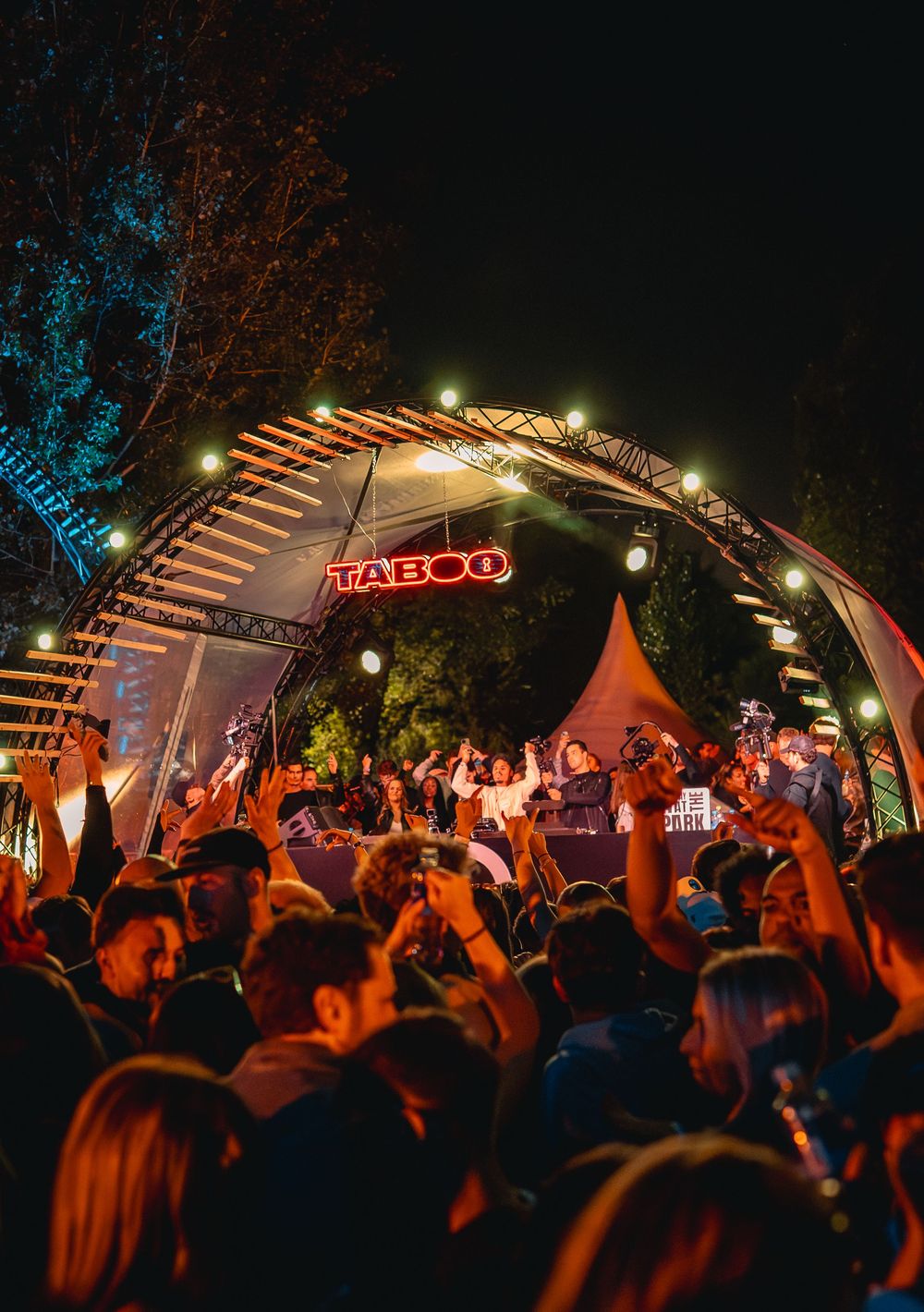Crowd enjoying a live outdoor music festival at night under a lit arch with a neon TABOO sign.