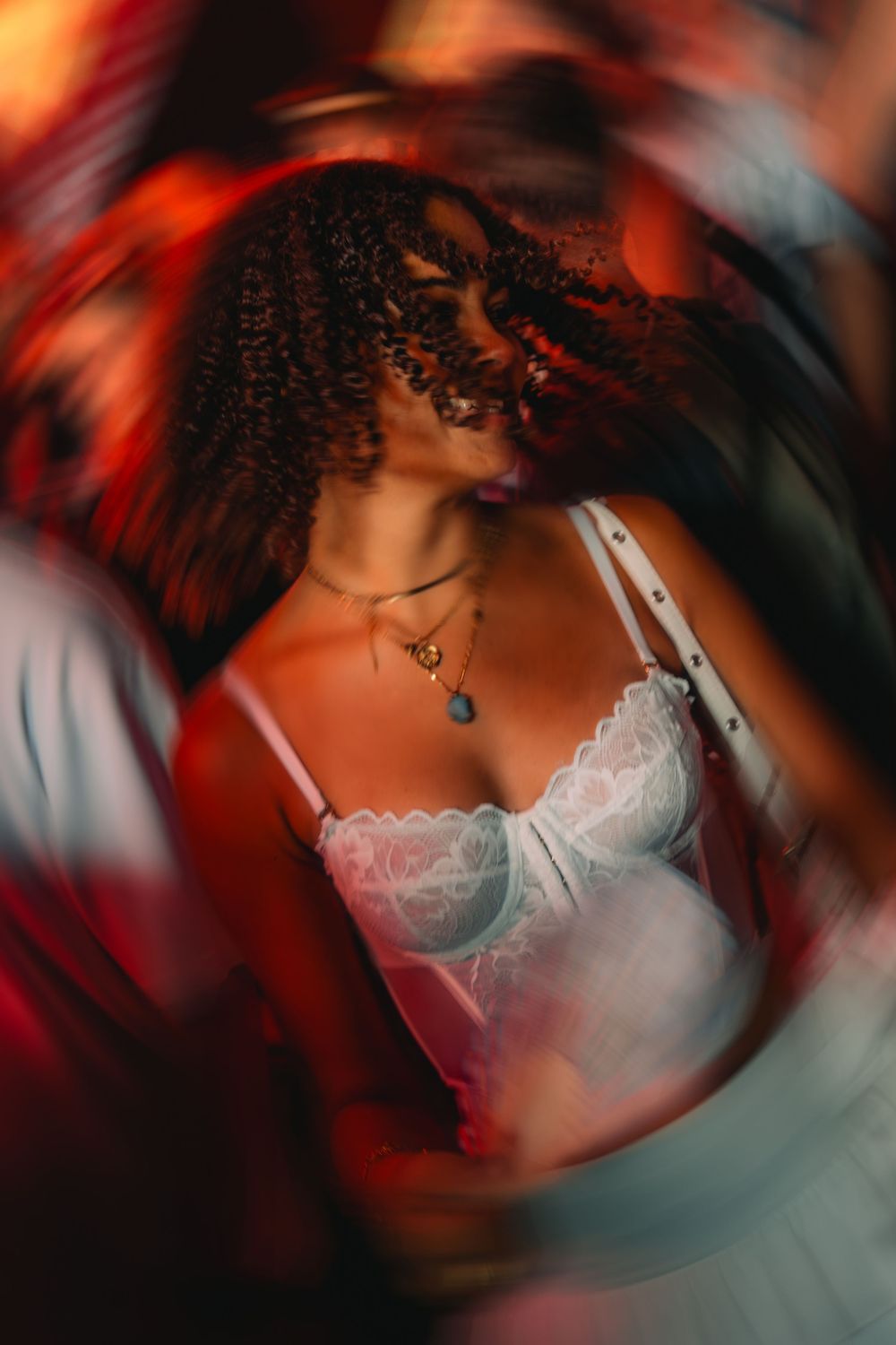 Young woman with curly hair dancing at a nightclub in a white lace top under red lights.